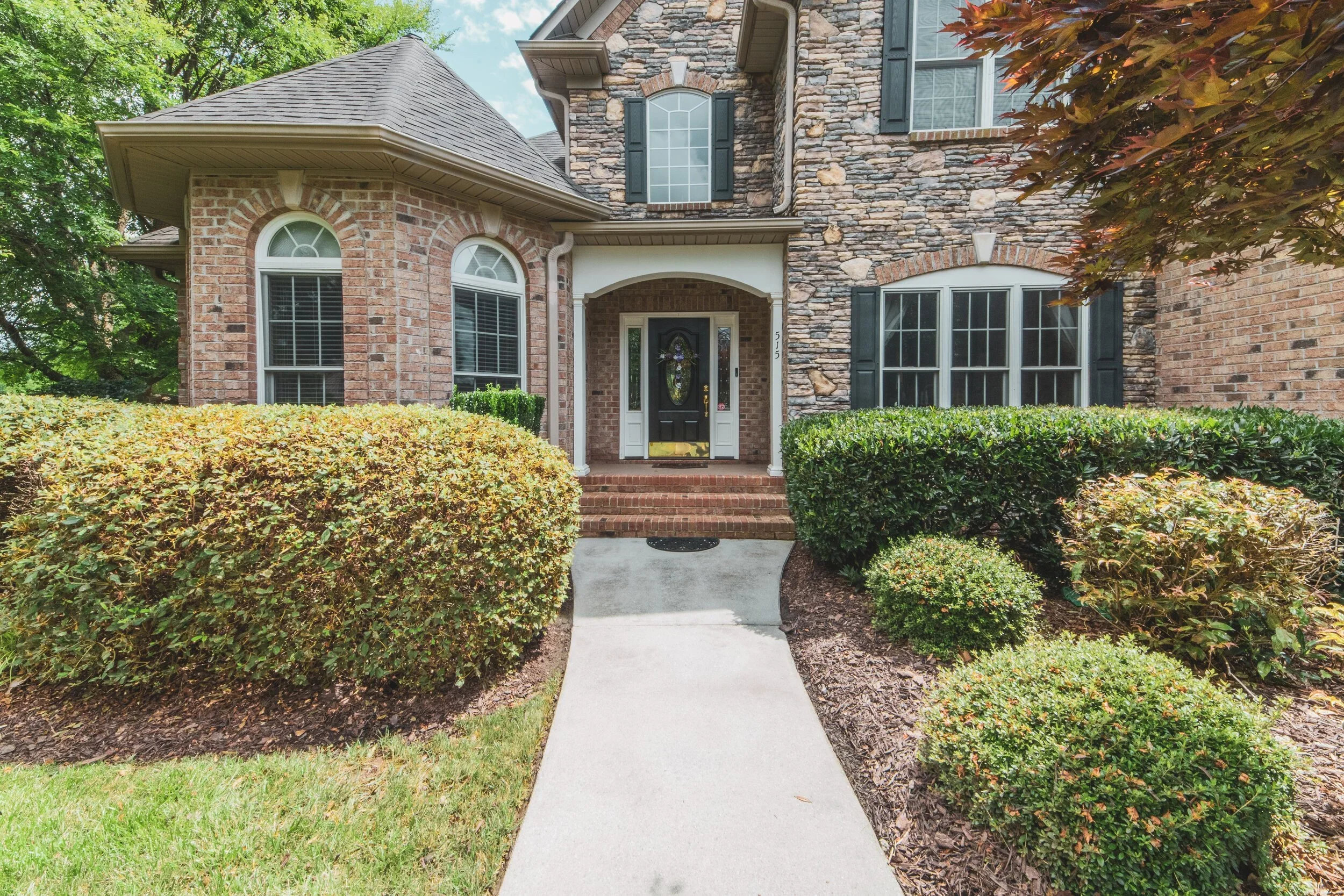 A brick and stone house with a black front door, surrounded by manicured bushes and trees, and a concrete walkway leading to the entrance.