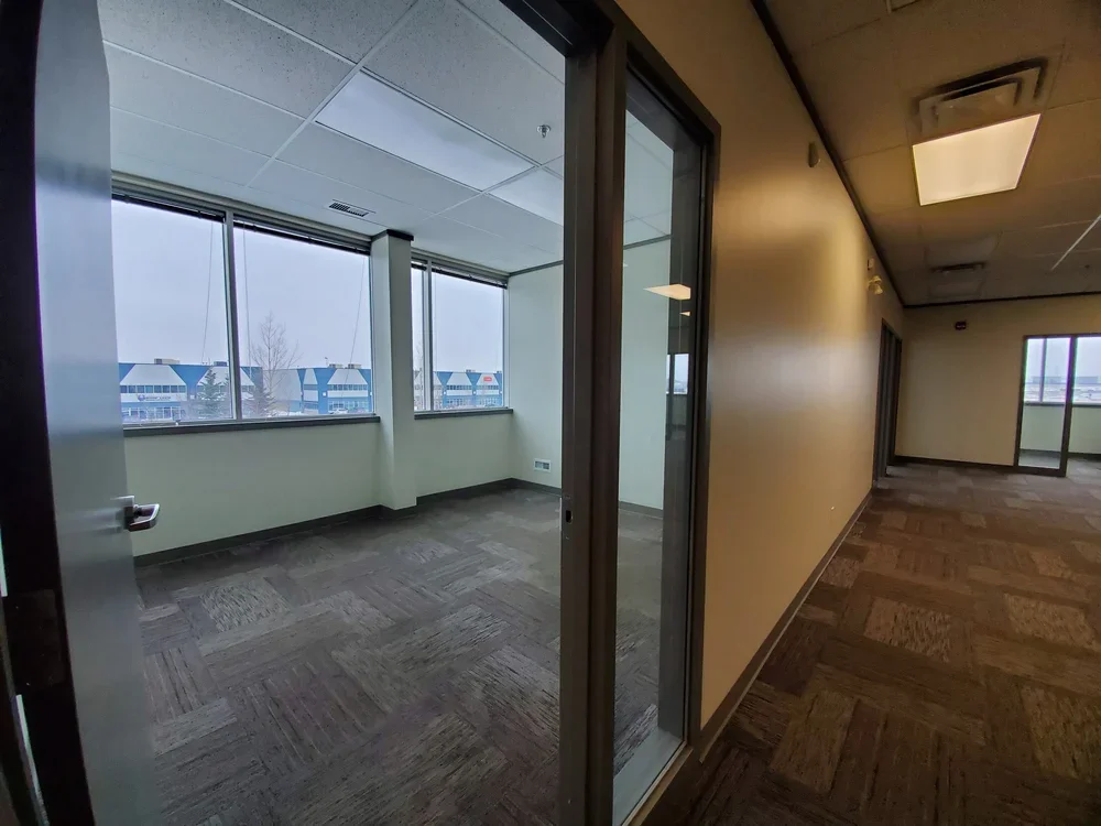 Interior of an office space with windows looking out onto an overcast day, with a small adjacent room visible through glass partitions, and empty with carpeted floors and ceiling lights.