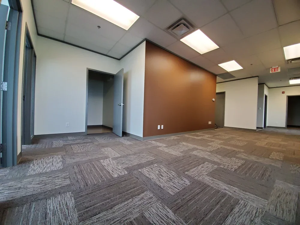Empty office space with carpeted floors, white and brown walls, ceiling lights, multiple doorways, and an exit sign.