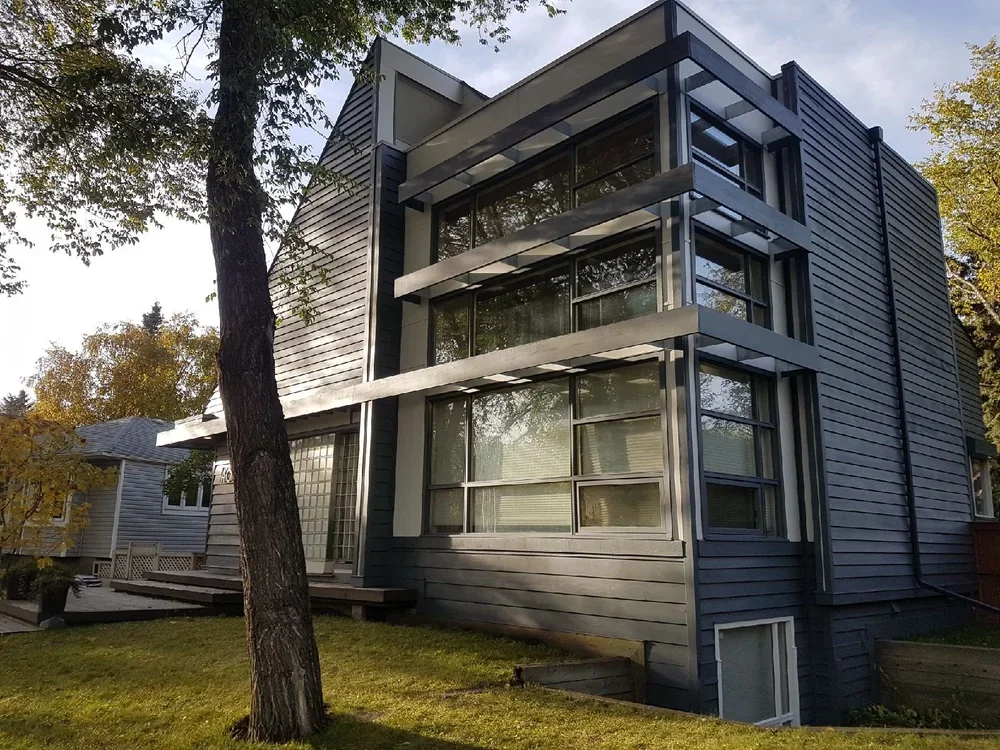 Modern three-story house with large glass windows and horizontal gray siding, surrounded by trees and a grassy yard.