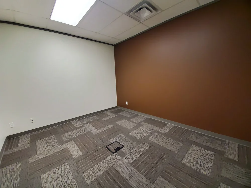 Empty office room with brown and white walls, carpeted floor, ceiling with fluorescent lights, and an air vent.