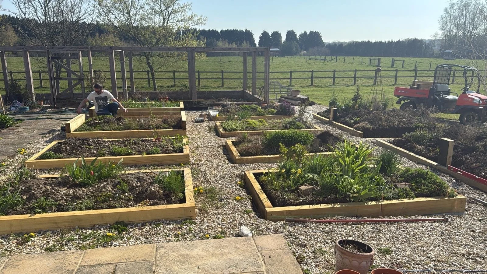 A person working in a garden with multiple rectangular garden beds on a gravel surface, surrounded by a rural landscape with green fields, trees, and a red utility vehicle.