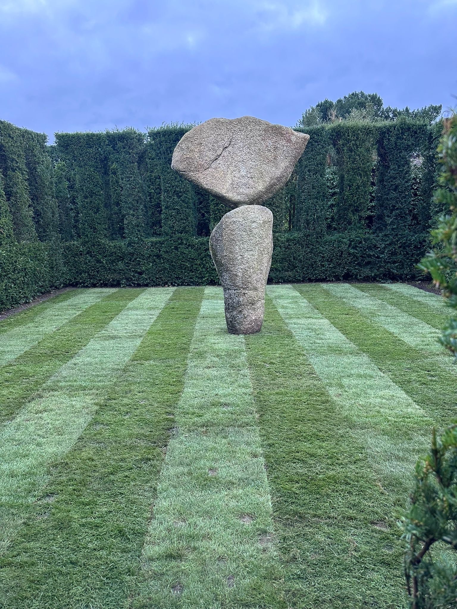 A stone sculpture with three large stones stacked vertically in a garden with neatly mowed grass and tall hedge in the background.