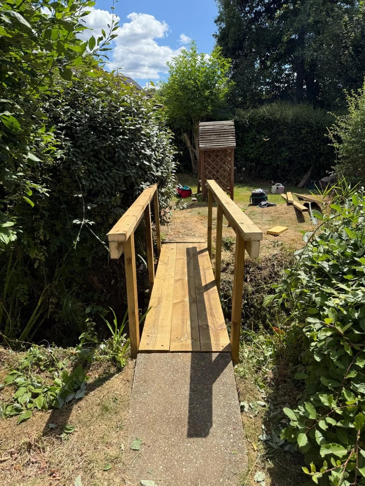 A small wooden bridge with handrails on both sides, leading to a yard with a wooden shed, picnic table, and various tools and objects on the ground, surrounded by greenery and bushes under a partly cloudy sky.