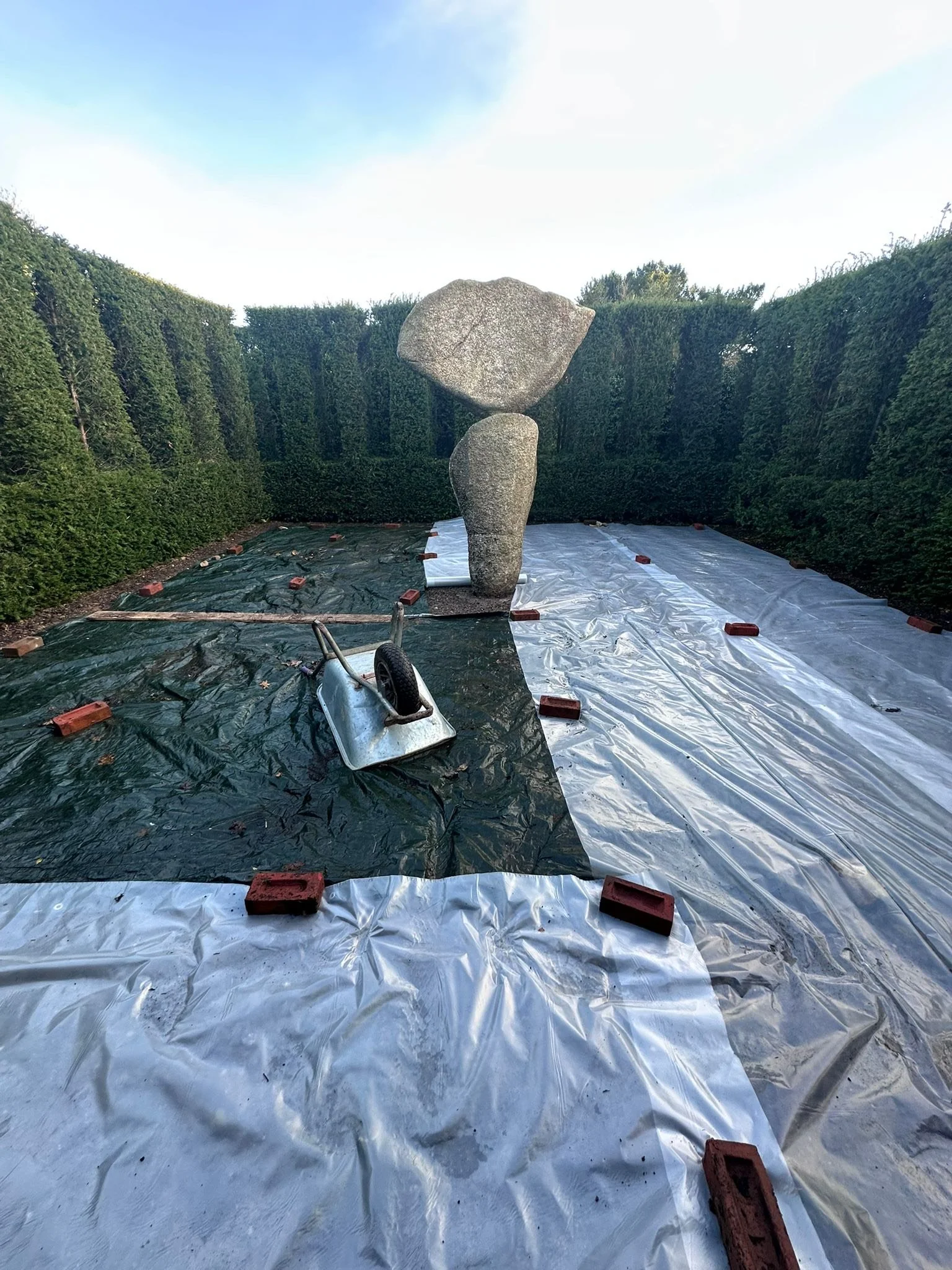 A garden with three large rocks balanced in a sculpture, surrounded by tall, trimmed hedges. The ground is covered with plastic sheets held down by bricks, with a wheelbarrow lying on its side nearby.
