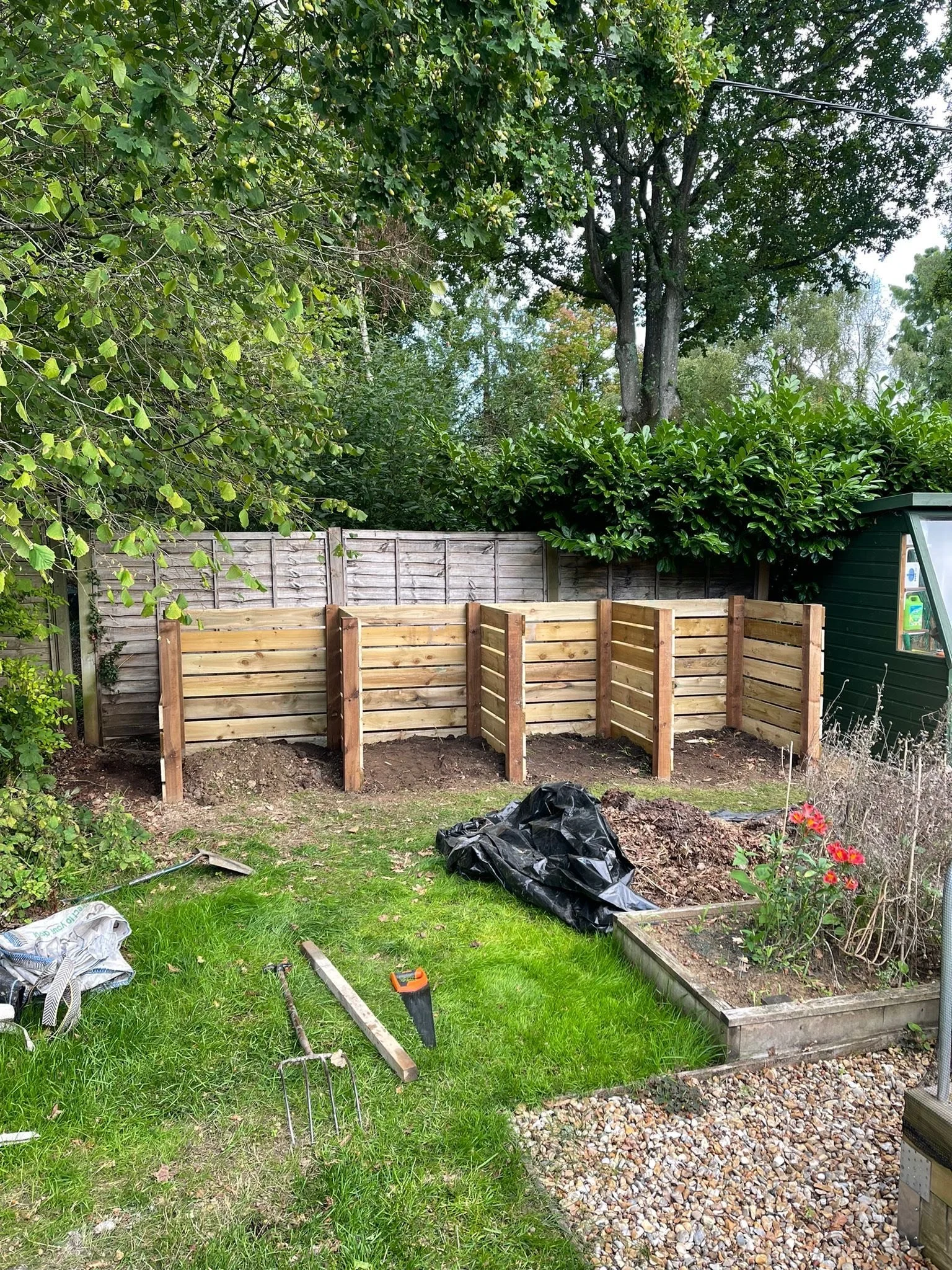 A backyard garden with a new wooden fence being built, various tools, a black trash bag, a flowerbed with red flowers, and trees in the background.