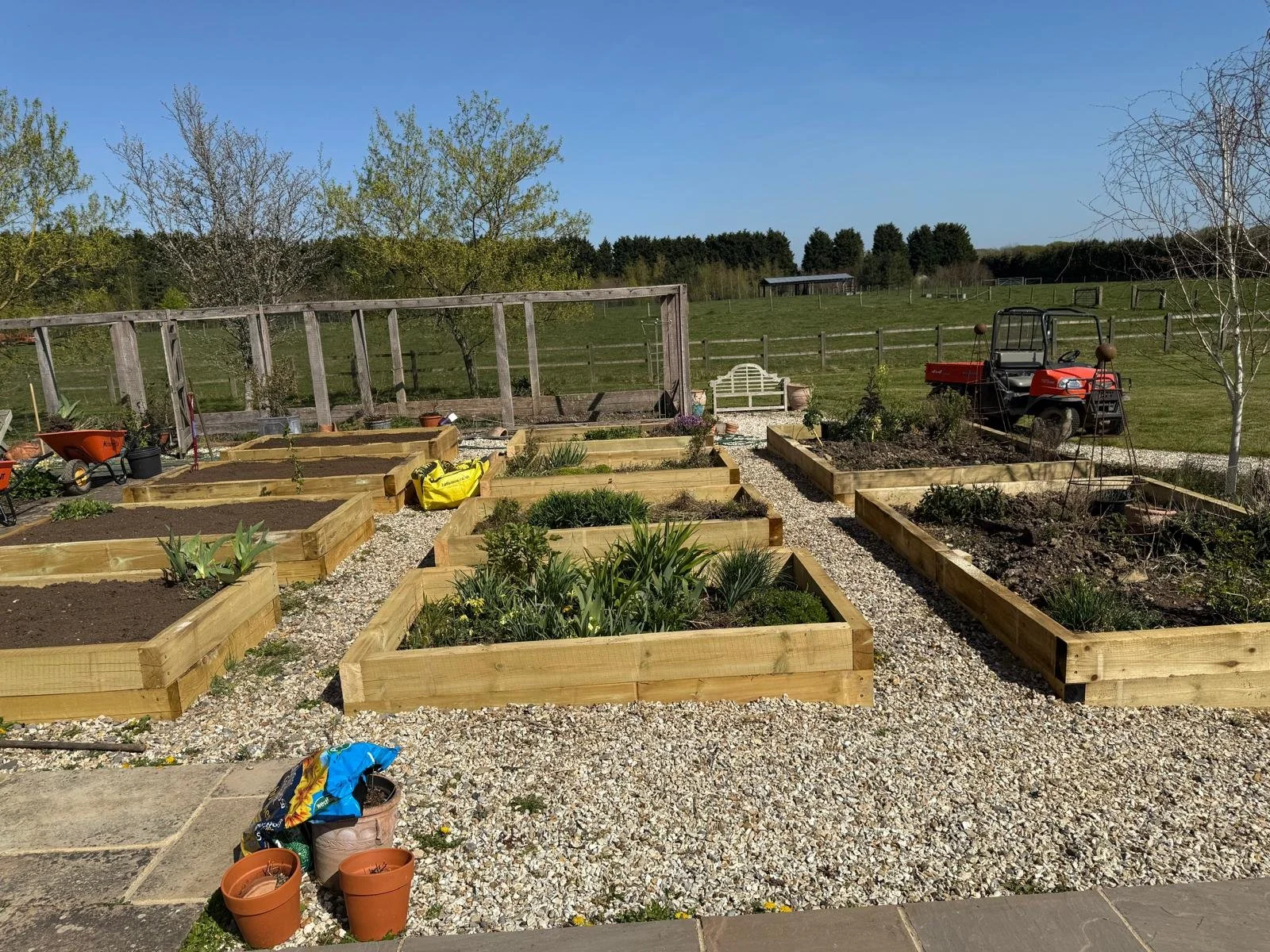 A garden with raised wooden beds filled with soil and growing plants, surrounded by gravel walkways and equipment, with a grassy field, trees, and blue sky in the background.