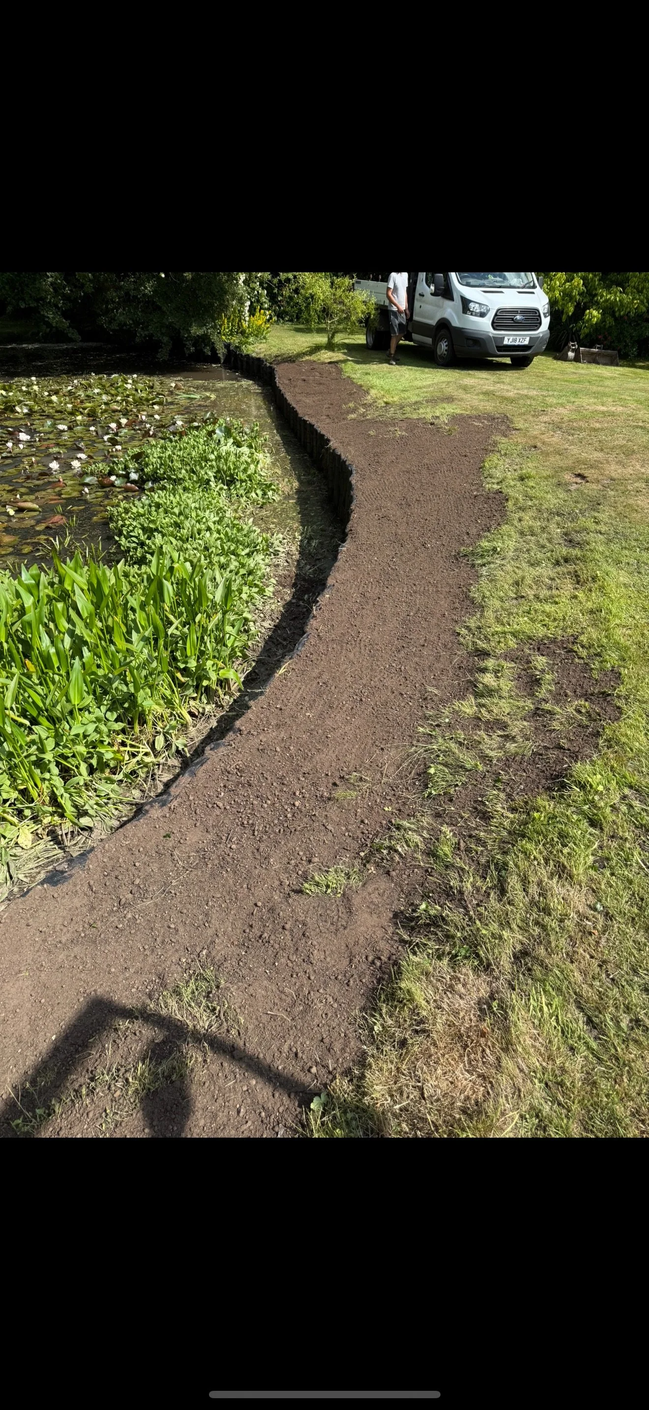 A newly laid dirt pathway along a pond with aquatic plants, next to a grassy lawn with a parked white van and a person standing nearby.