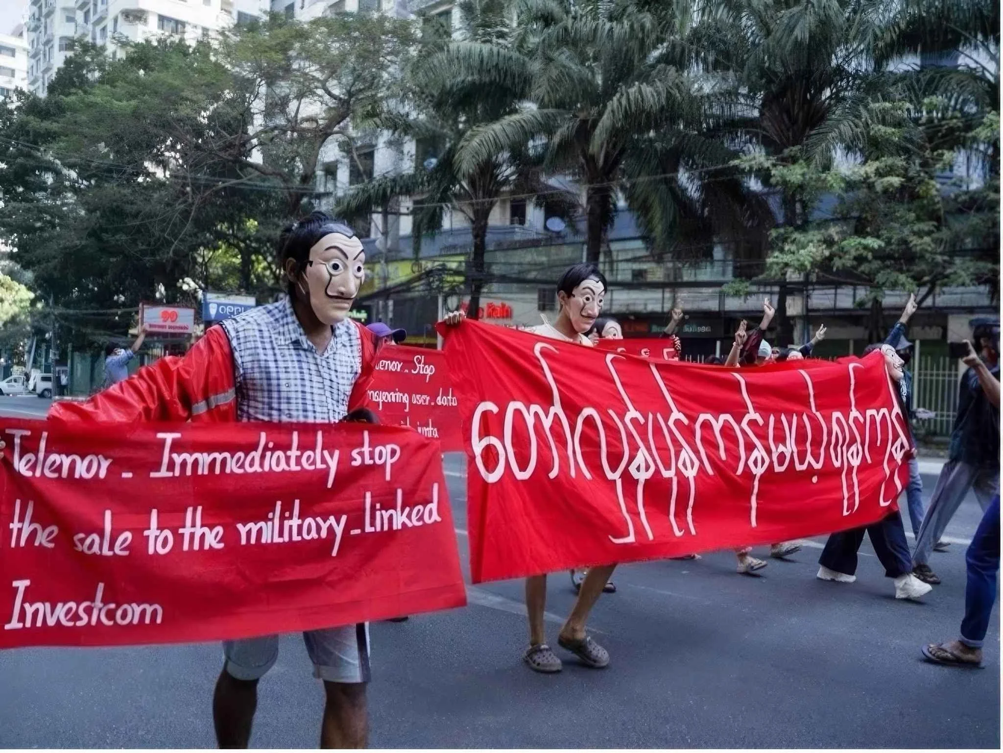 Personer iført masker som forestiller politiske ledere, som deltar i en demonstrasjon med store røde bannere med tekst. Demonstrasjonen foregår på gaten med trær og bygninger i bakgrunnen.