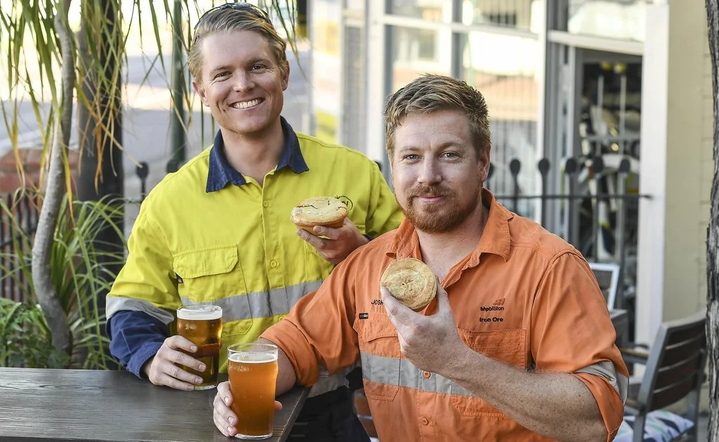 Two men sitting outside at a table, smiling, holding pastries and beers, with plants and a house in the background.