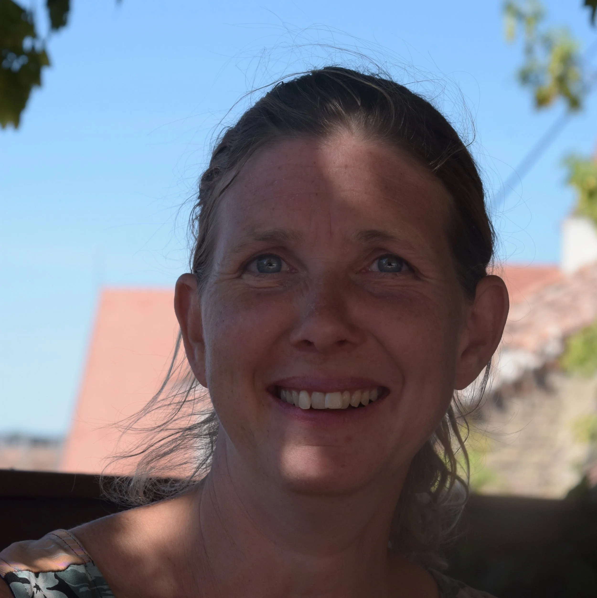 Mit Sonnenlicht beschattetes Gesicht einer lachenden Frau, blauer Himmel und bunte Gebäude im Hintergrund.