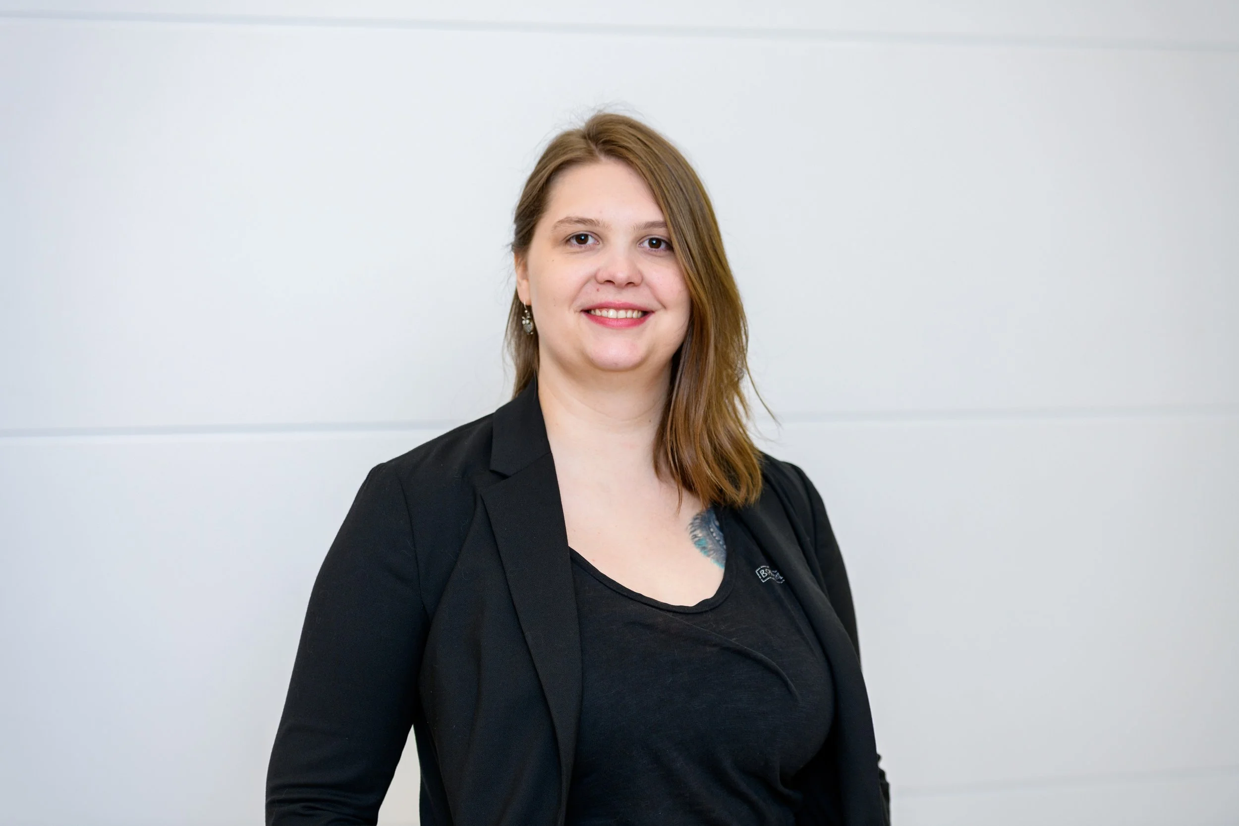 Frau mit braunen Haaren, schwarze Jacke, lächelnd vor weißer Wand.
