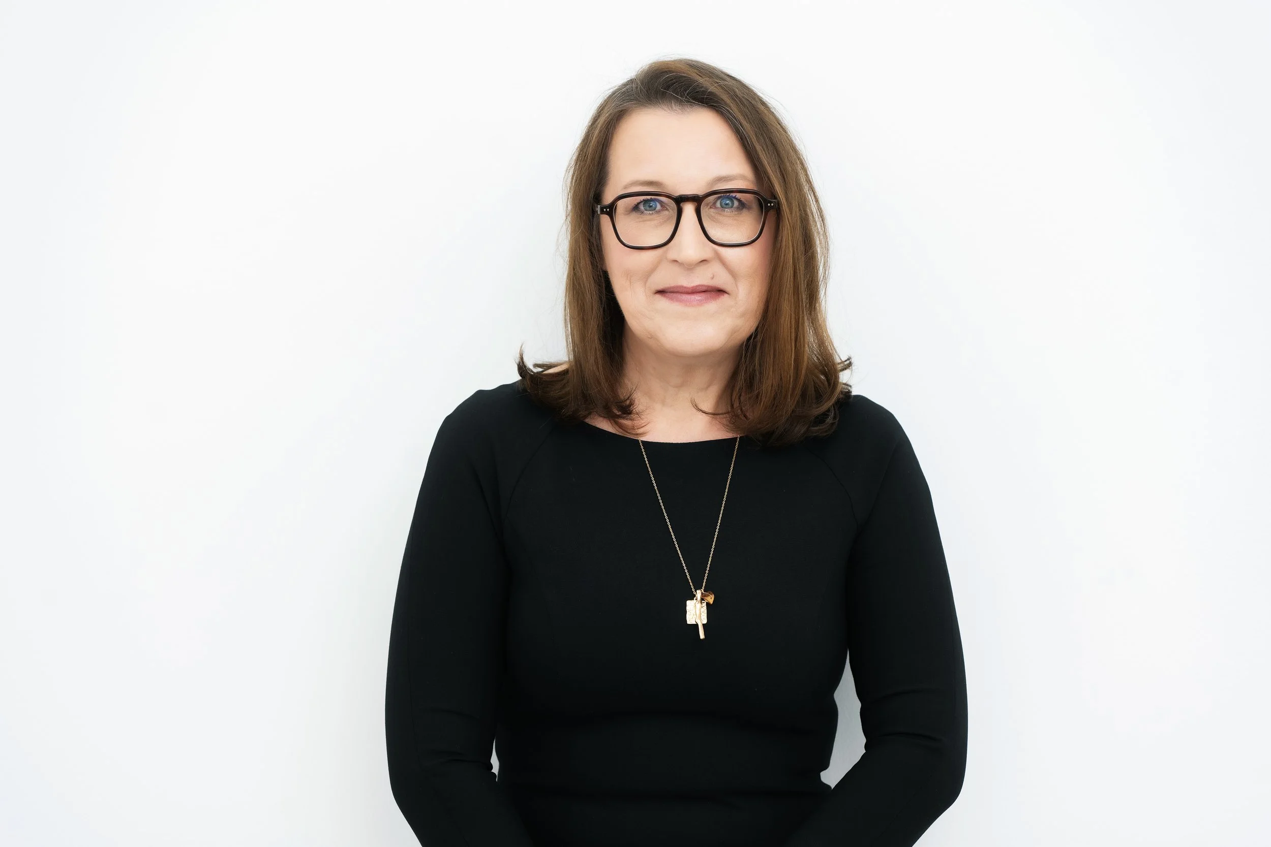 Eine Frau mit braunen Haaren, schwarzen Brillen und schwarzem Oberteil vor weißem Hintergrund.