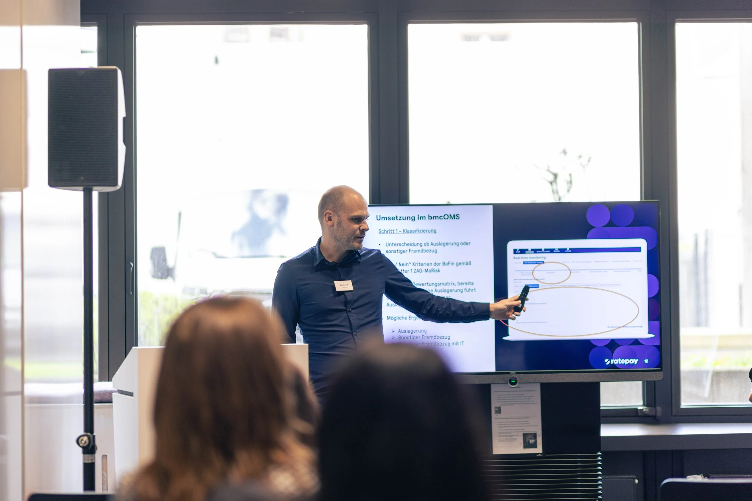 Ein Mann hält ein Smartphone in der Hand, während er in einem Konferenzraum vor einem Bildschirm präsentiert. Der Bildschirm zeigt eine Präsentation mit Texten und Diagrammen, und es gibt mehrere Teilnehmer im Raum.