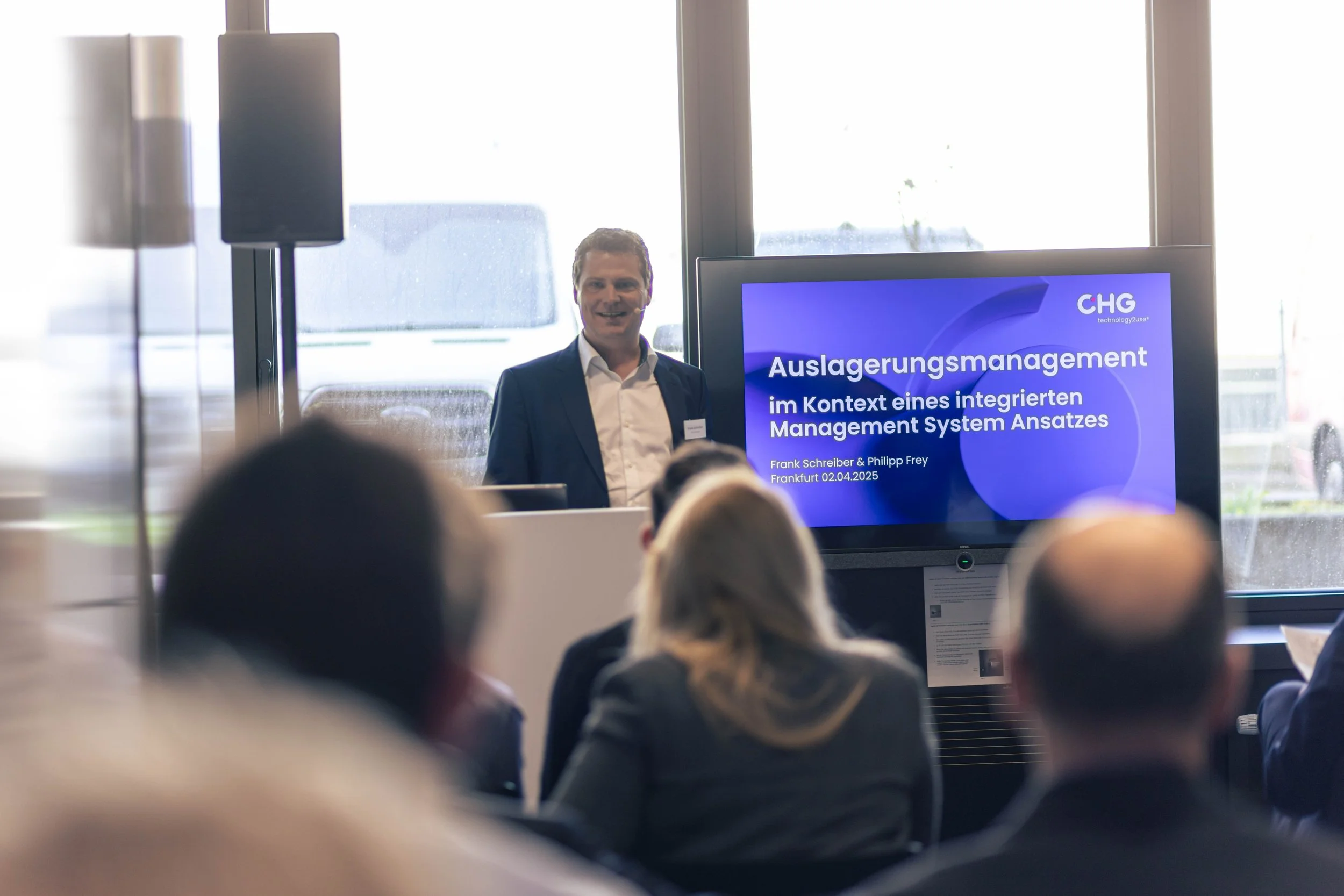 Ein Mann in Anzug hält eine Präsentation vor Publikum bei einem Kongress, auf einem Bildschirm steht der Titel "Auslagerungsmanagement im Kontext eines integrierten Management System Ansatzes".