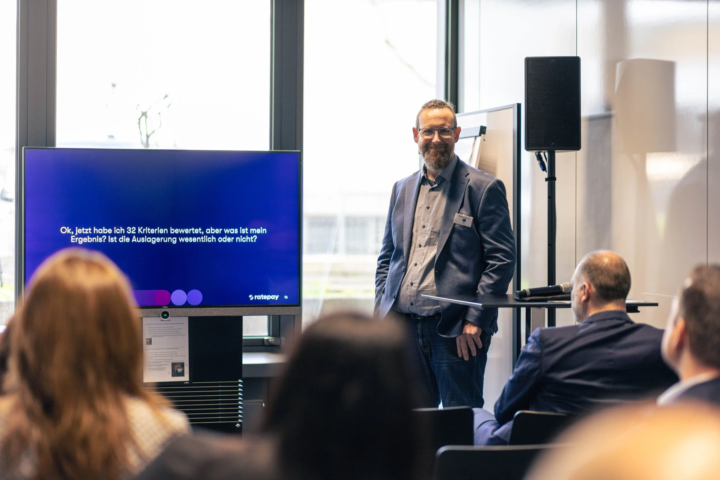 Ein Mann hält eine Präsentation vor einem Publikum in einem modernen Raum mit großen Fenstern. Auf dem Bildschirm ist Text auf Deutsch sichtbar.