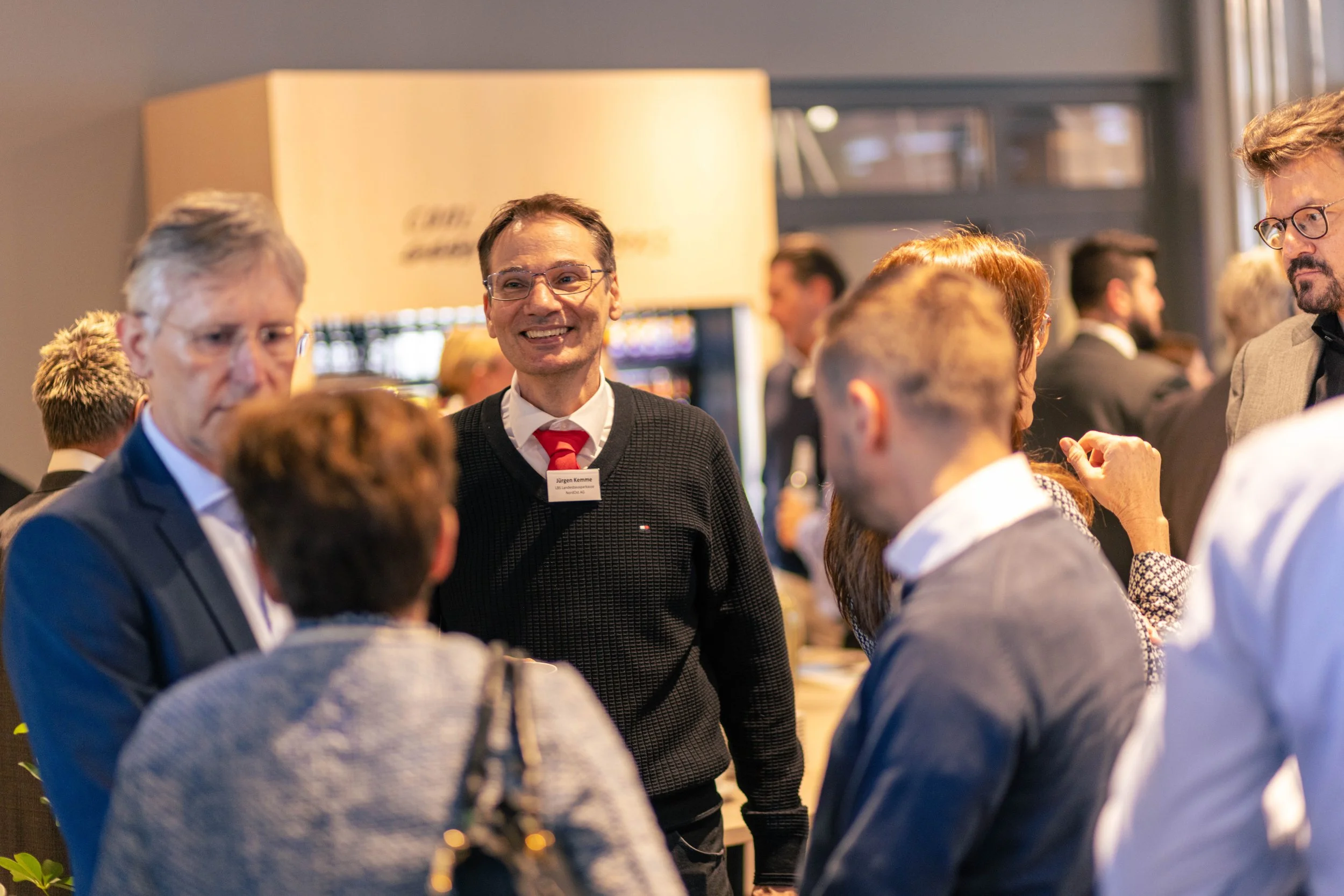Menschen bei einer Konferenz im Gespräch, ein Mann mit roten Krawatte lächelt, alle tragen formelle Kleidung.