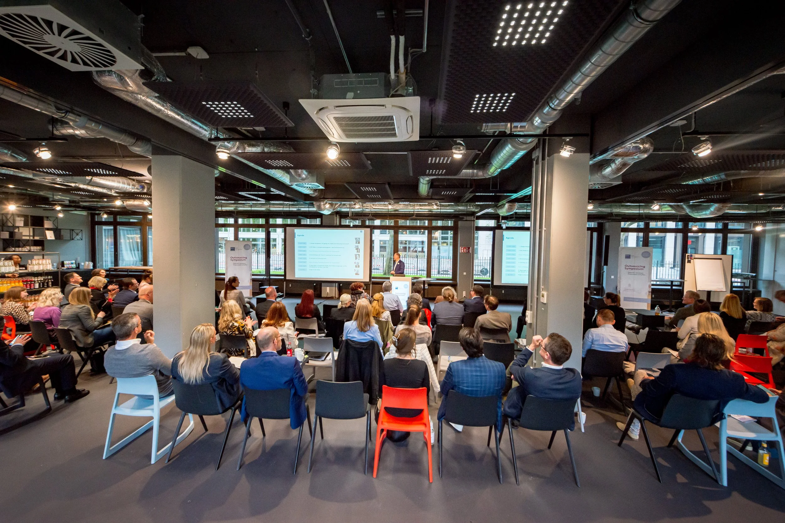 Eine große Gruppe von Menschen sitzt in einem modernen Konferenzraum, während ein Redner an einem Podium vor Präsentationsbildschirmen spricht.