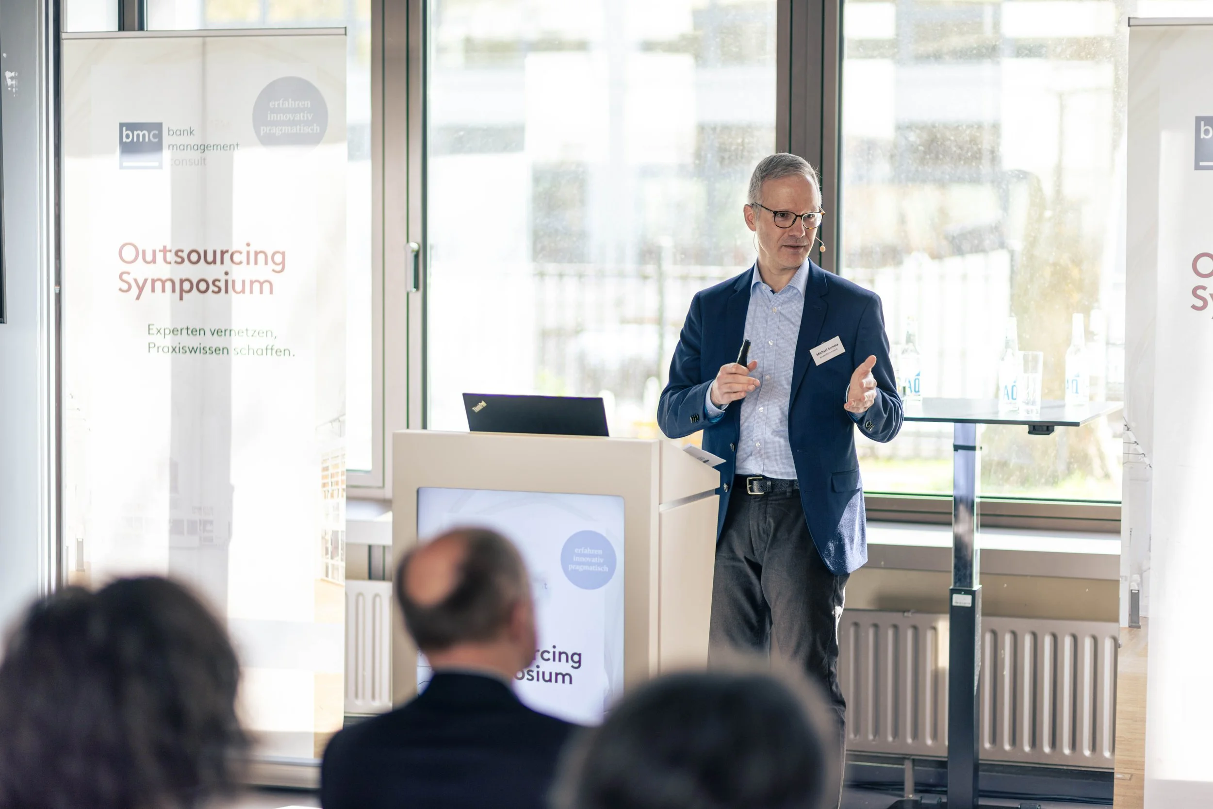 Ein Mann hält eine Präsentation bei einem Outsourcing-Symposium, im Hintergrund Banner mit dem Text 'Outsourcing Symposium' und Deutschlandkarte, vorne im Publikum sitzen wenige Personen.