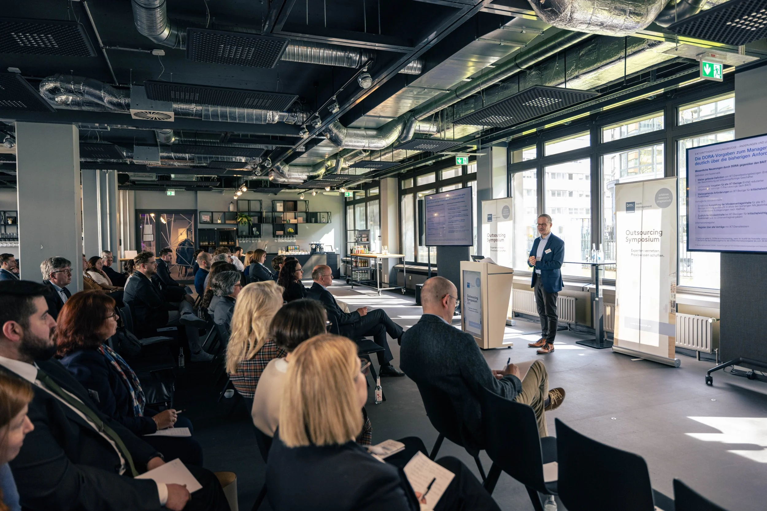 Ein Business-Meeting oder eine Konferenz mit vielen Teilnehmern, die einem Präsentationsvortrag zuhören, in einem modernen Raum mit großen Fenstern und Bildschirm.