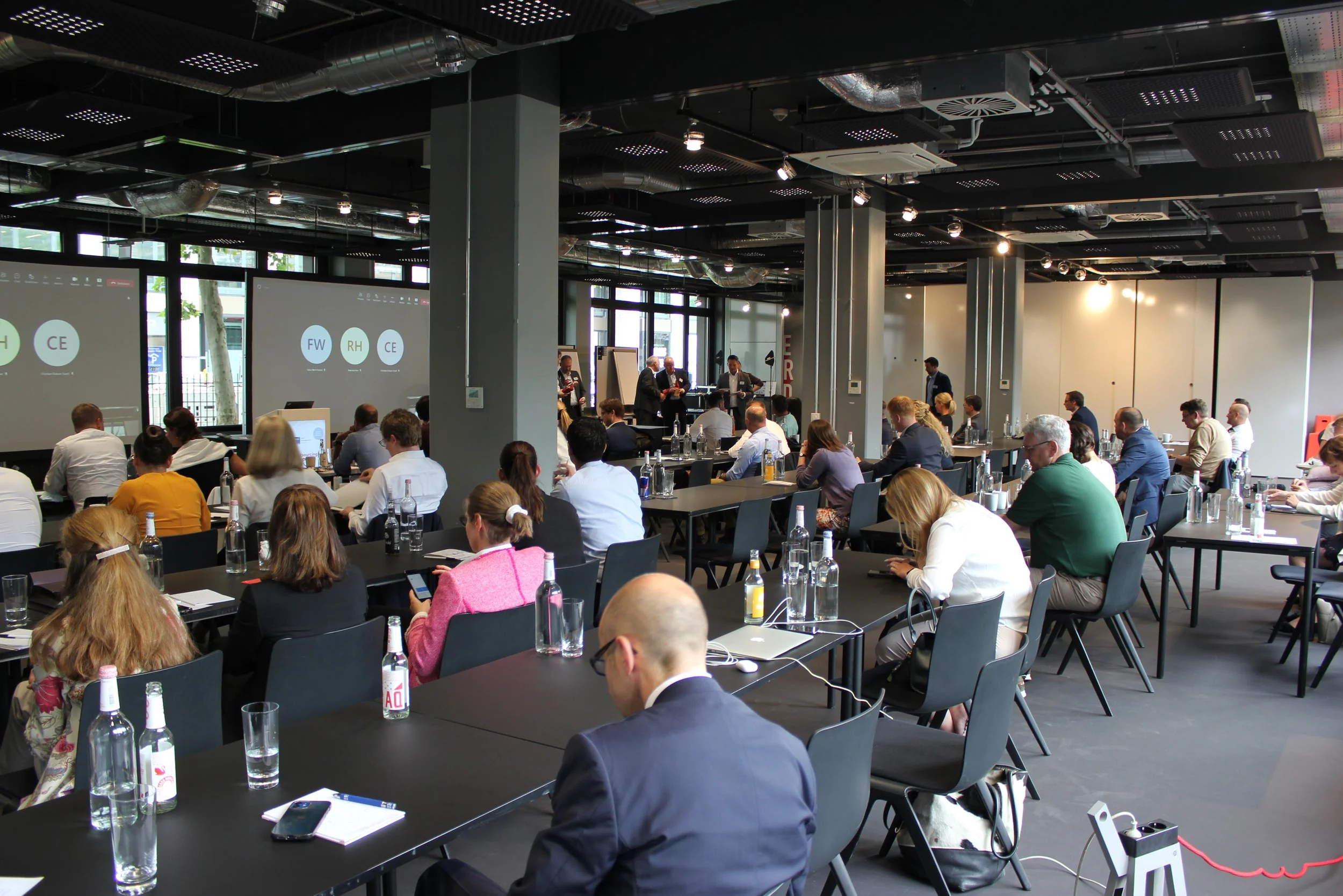 Innenaufnahme eines Konferenzraums während eines Seminars oder Workshops mit vielen Teilnehmern, Projektionsbildschirme an der Wand, die Präsentationen zeigen, und Moderatorengruppe vorne bei der Diskussion.