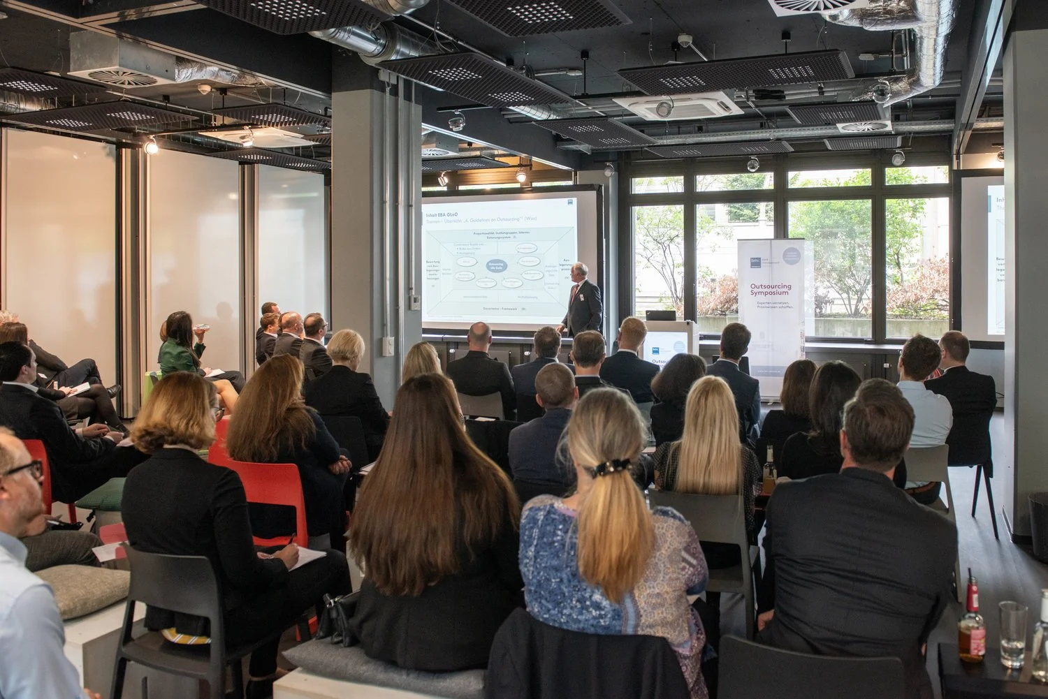 Ein Vortrag während eines Outsourcing-Symposiums in einem modernen Konferenzraum mit vielen Zuschauern, großen Fenstern und einem Präsentationsbildschirm.