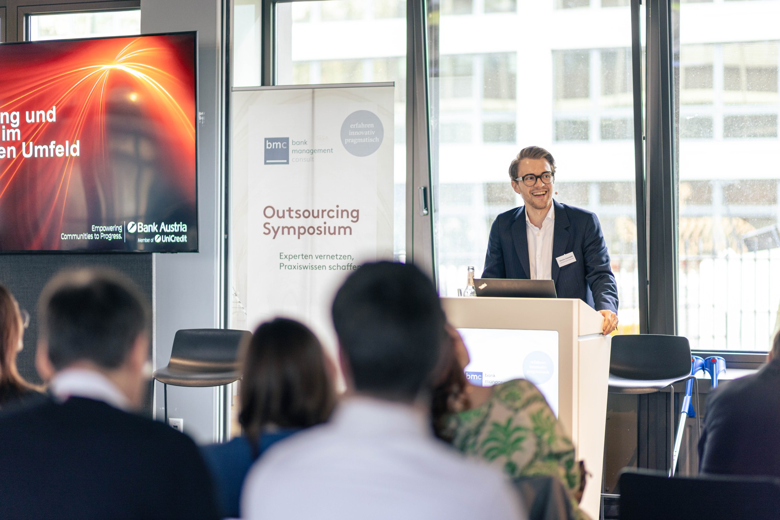 Ein Mann steht an einem Podium und hält eine Präsentation bei einer Outsourcing-Symposium, im Hintergrund sind Schilder und ein Bildschirm mit Produkten- und Unternehmensinformationen sichtbar. Im Publikum sitzen mehrere Personen und hören zu.