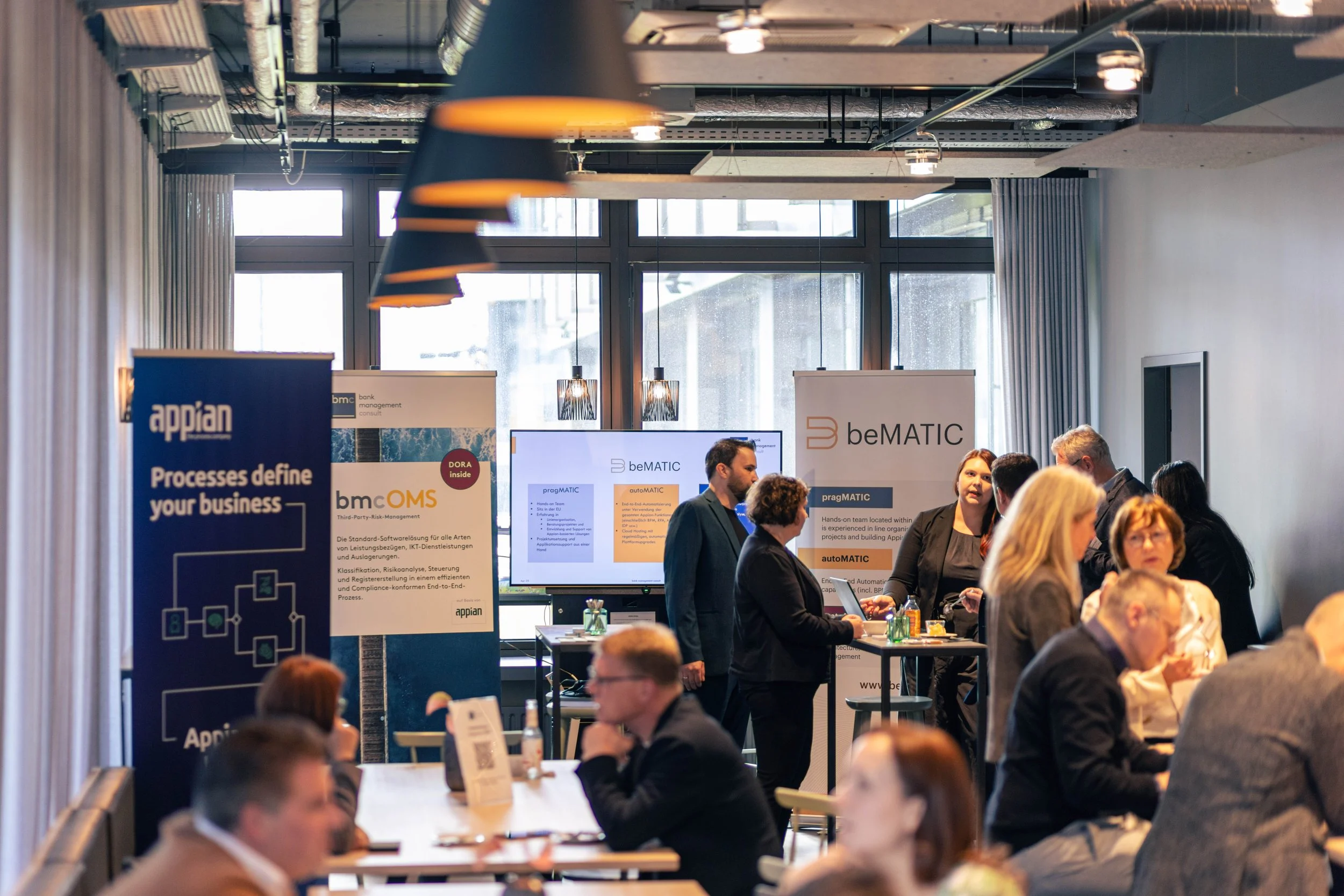 Geschäftsleute bei einer Konferenz oder Messe, Gespräche vor Informationsständen, Banner mit Firmennamen und Präsentationsdisplays im Hintergrund.