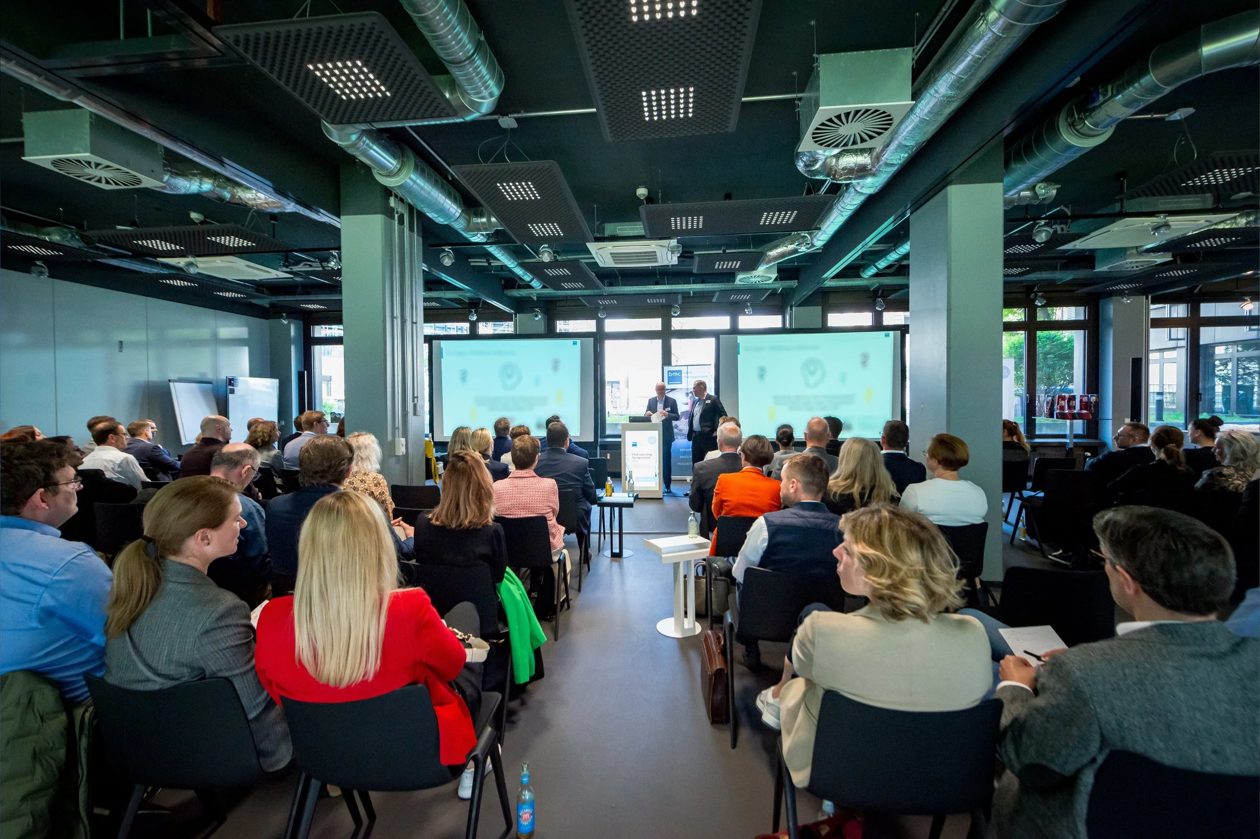 Ein Business-Meeting mit vielen Menschen in einem modernen Konferenzraum, zwei Präsentationsbildschirme, drei Redner vor Publikum, große Fenster, technische Decke mit Lüftungsrohren.