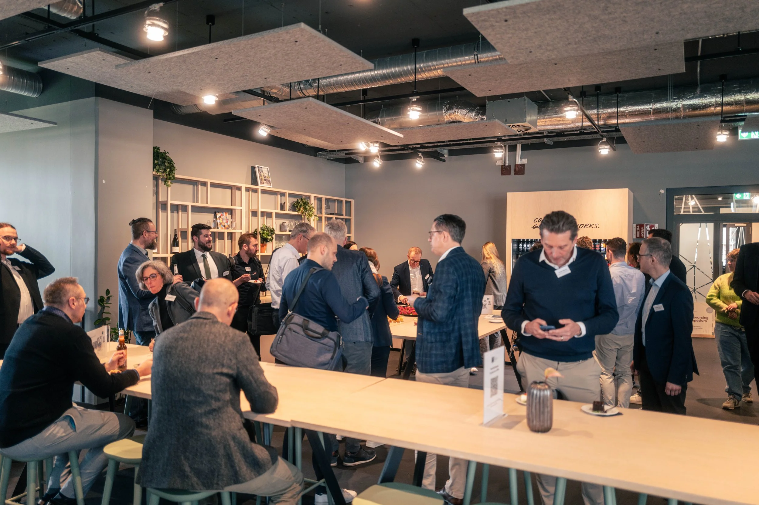 Menschen bei einem Networking-Event in einem modernen Raum mit Stehtischen, einige sitzen, andere stehen und sprechen, eine Deko-Regal im Hintergrund.