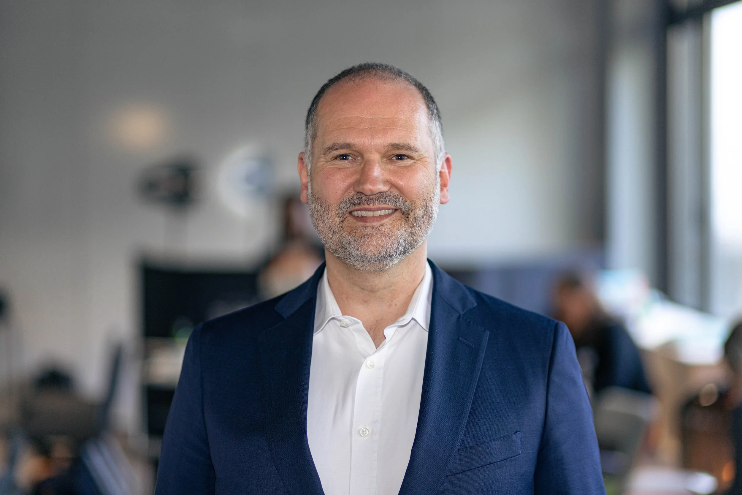 Ein lächelnder Mann in einem dunkelblauen Anzug und weißem Hemd, im Büro mit anderen Mitarbeitern im Hintergrund.