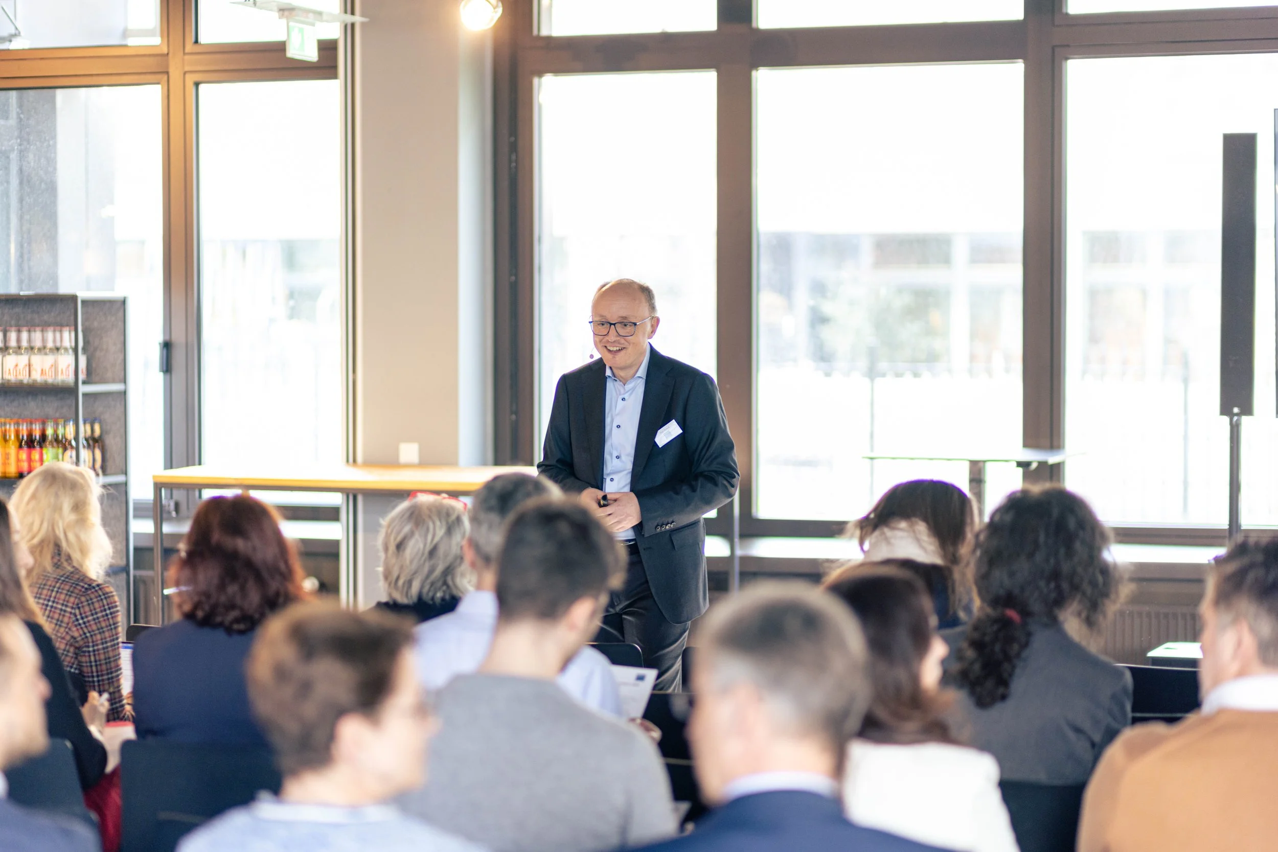 Ein Mann hält einen Vortrag vor einem Publikum in einem gut beleuchteten Raum mit großen Fenstern im Hintergrund.