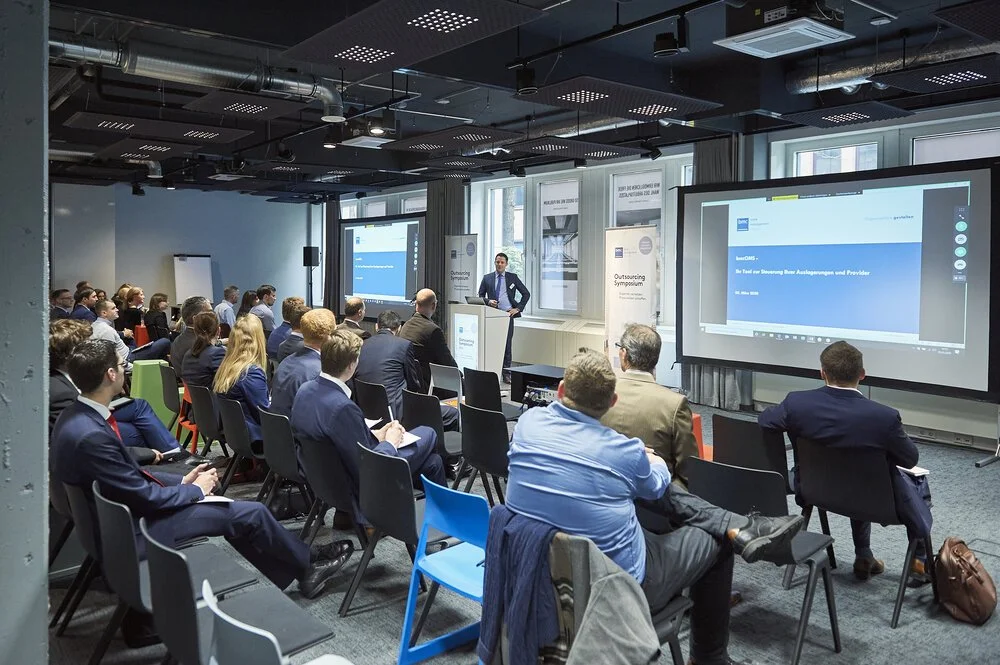 Ein Konferenzraum mit vielen sesselnden Menschen, die einem Vortrag zuhören. Es gibt zwei große Bildschirme mit Präsentationen. Der Raum hat große Fenster und moderne Dekoration.
