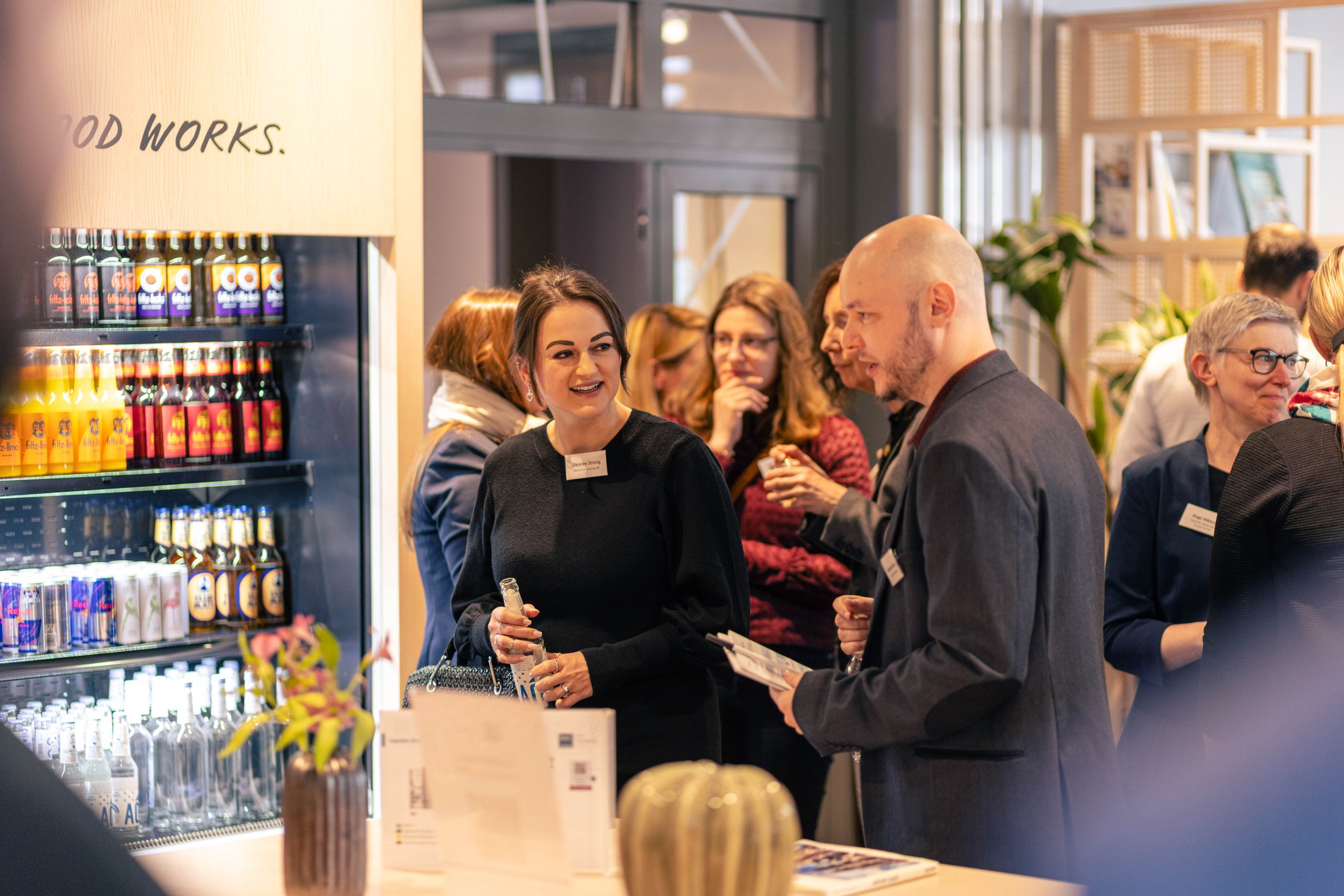 Menschen bei einer Networking-Veranstaltung, sprechen und trinken Getränke, im Hintergrund Getränke Kühlschrank mit Softdrinks, moderne helle Inneneinrichtung.