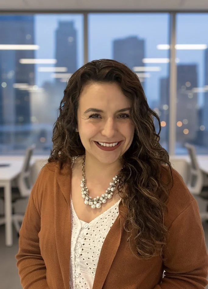 Lächelnde Frau mit lockigem braunem Haar, braunem Blazer und Perlenkette vor einem Bürofenster mit Stadthintergrund.