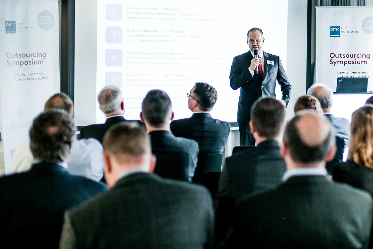 Ein Mann in Anzug spricht während eines Symposiums vor einem Publikum von Geschäftsleuten. Der Raum ist professionell eingerichtet, mit einem großen Bildschirm im Hintergrund, der eine Präsentation zeigt.