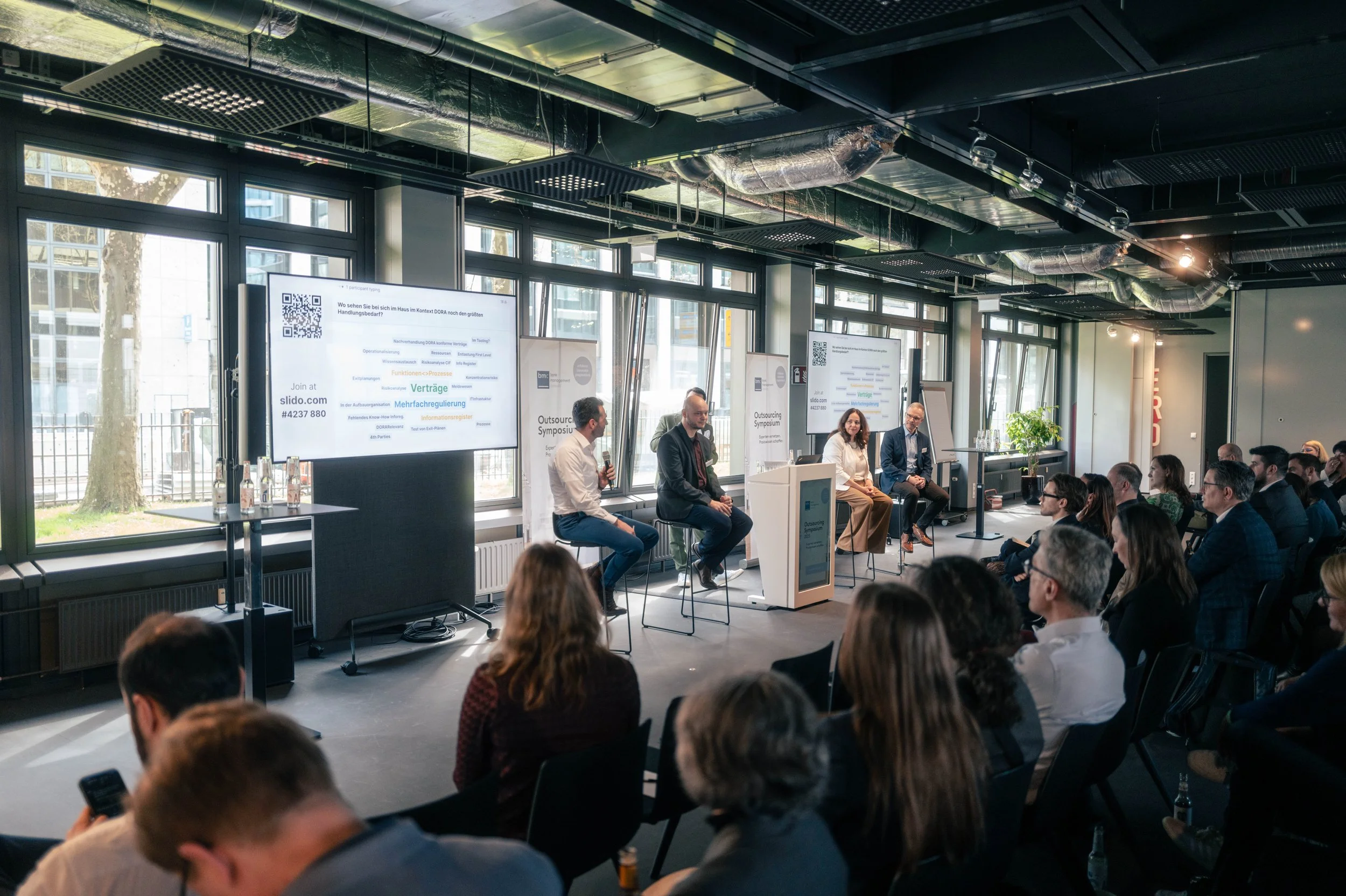 Paneldiskussion bei einer Konferenz mit fünf Sprechern auf der Bühne und einem Publikum, moderne Räumlichkeiten mit großen Fenstern und sichtbarer Decke.