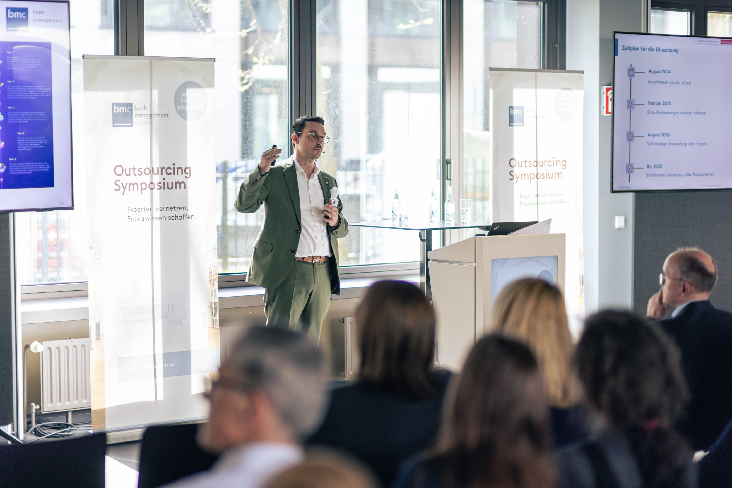 Ein Mann hält eine Präsentation auf einer Konferenz mit dem Titel "Outsourcing Symposium". Er steht vor einem Bildschirm und spricht zu einem Publikum, das im Raum sitzt.