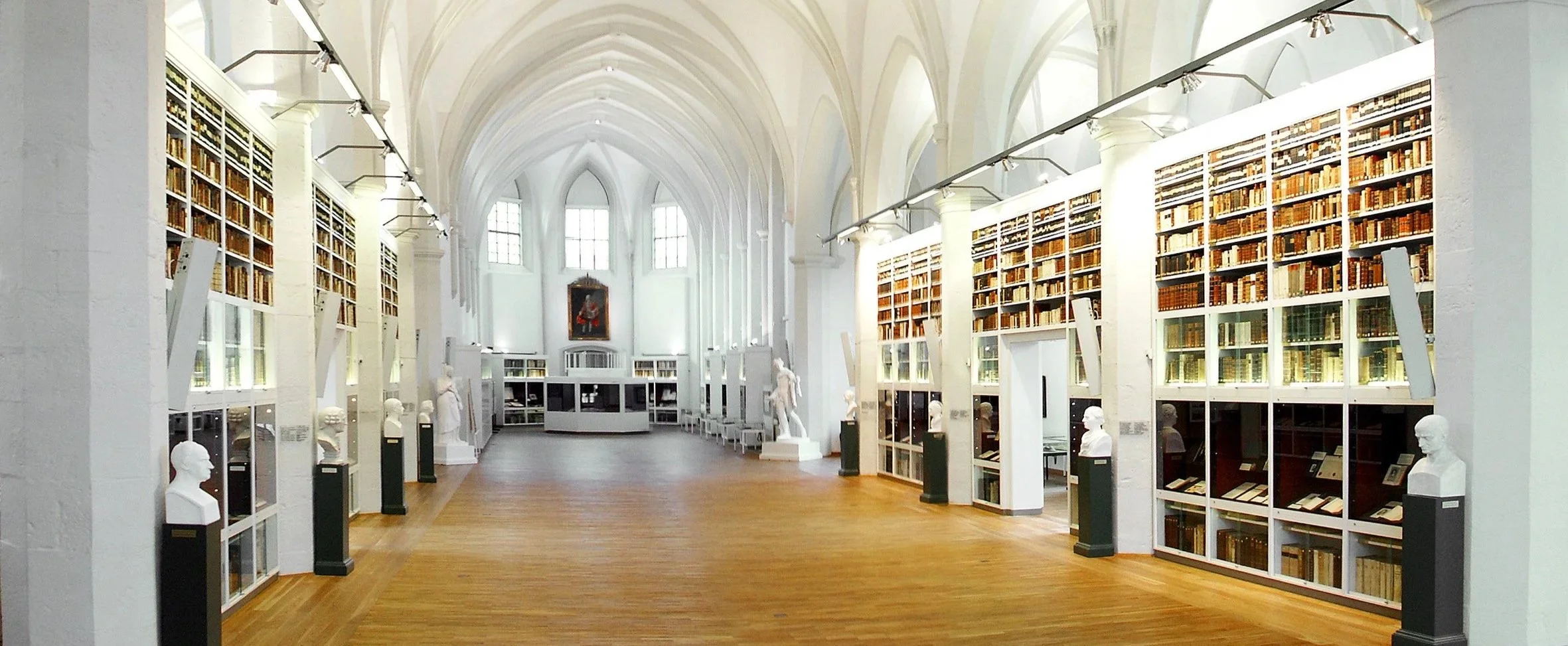 Blick in eine große, helle Bibliothek mit hohen, weißen Wänden, großen Fenstern und Bücherregalen mit alten Büchern. Statues und Informationstafeln sind auf Podesten platziert, das Holzparkett bedeckt den Boden.