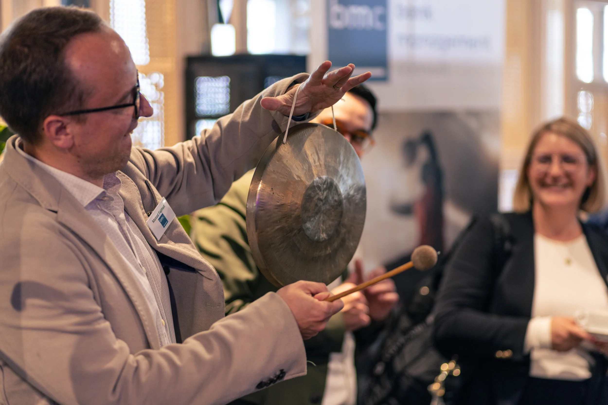 Mann mit Brille spielt mit einer Gongscheibe während einer Veranstaltung, im Hintergrund Menschen die lachen und einen Banner.
