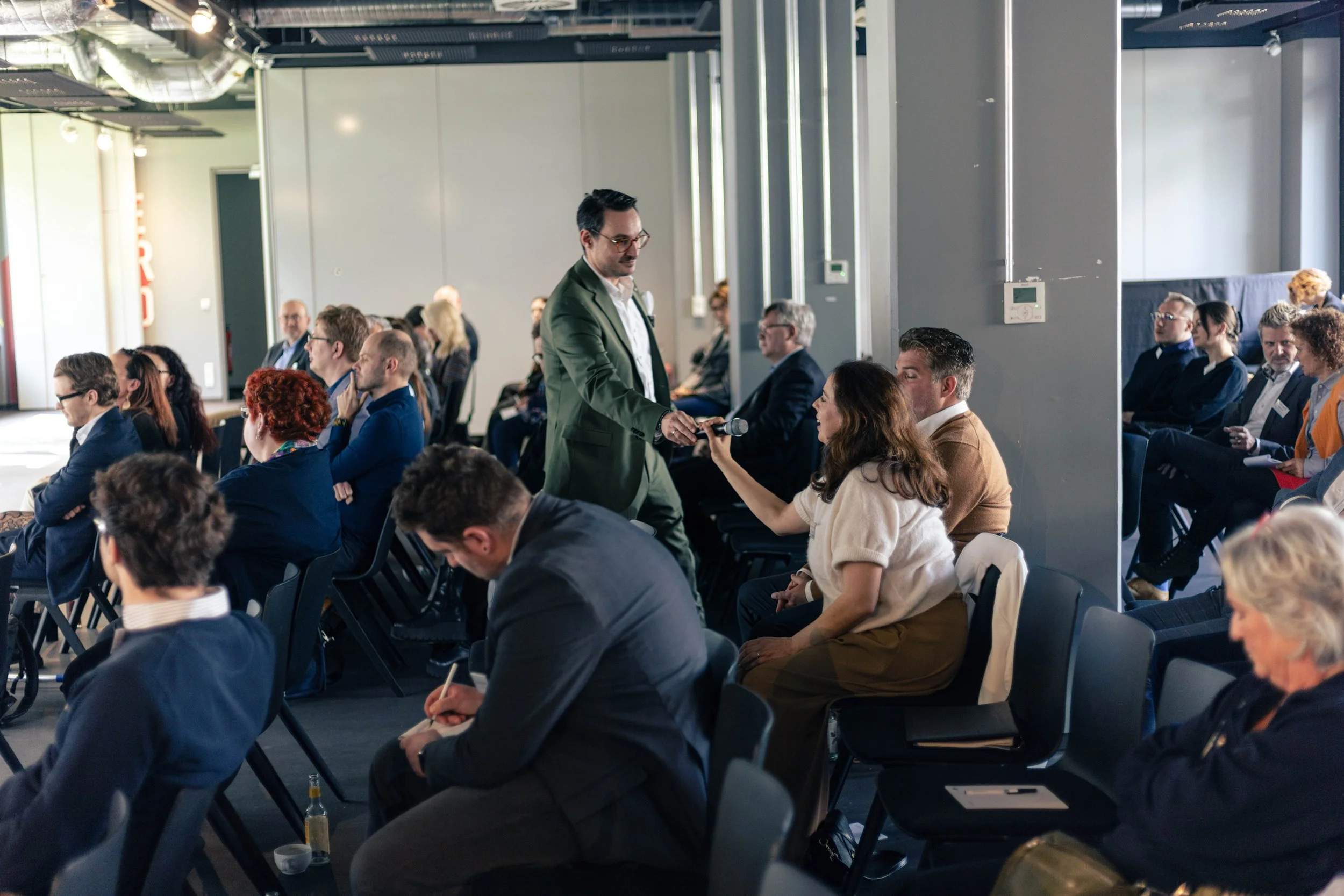 Ein Mann in einem grünen Anzug gibt einem weiblichen Sprecher ein Mikrofon in einem Konferenzraum mit vielen Sitzplätzen.
