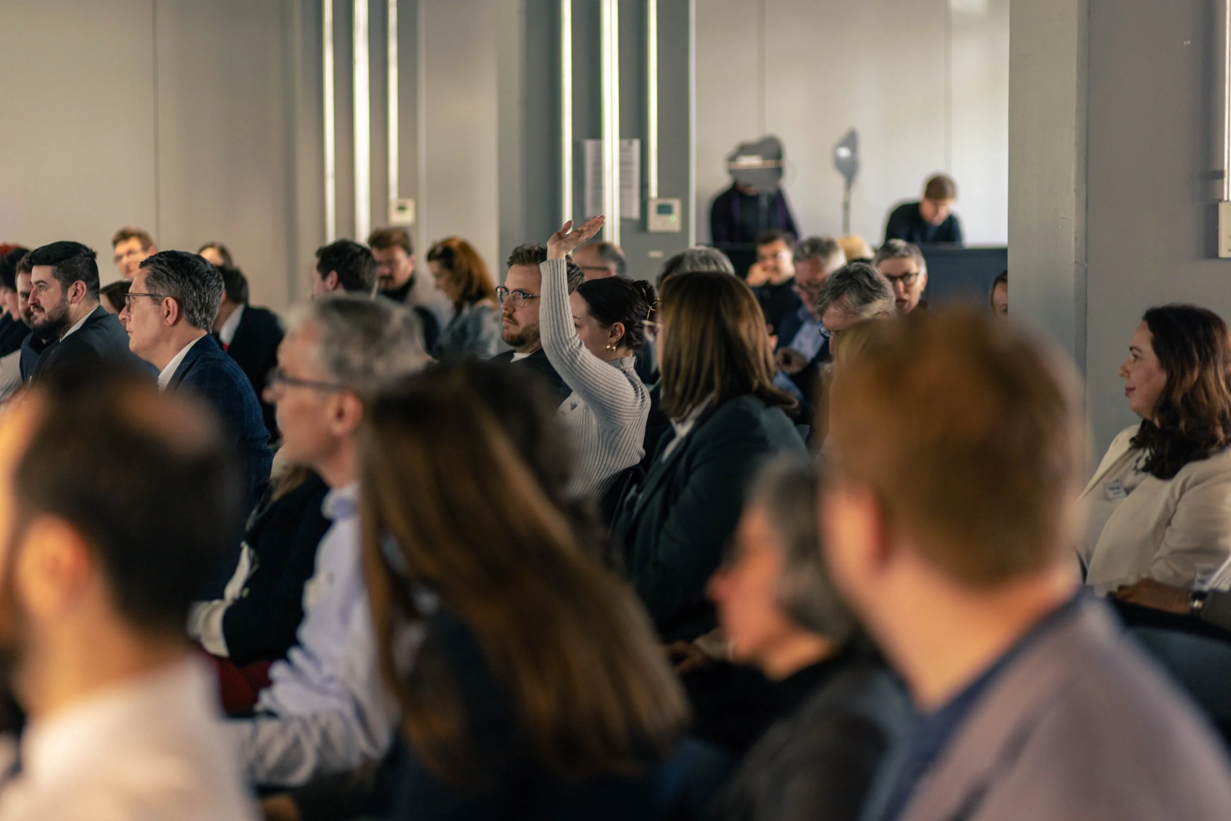 Menschen sitzen in einem Conference-Raum, eine Frau hebt die Hand, während andere zuhören.