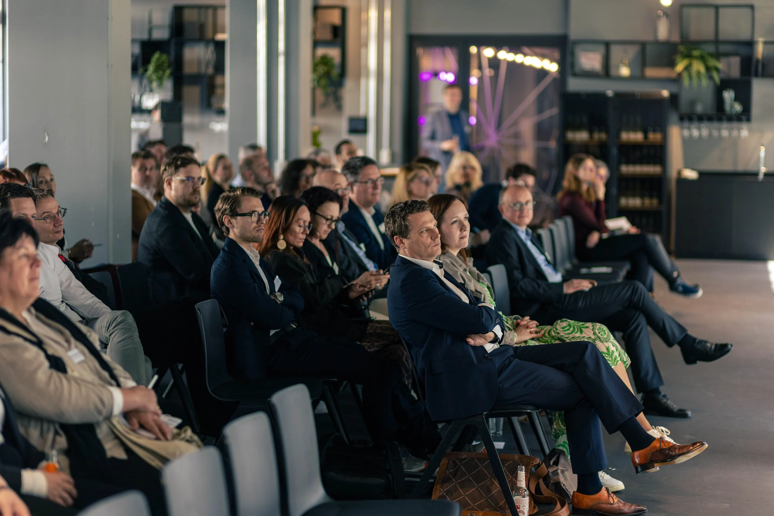 Menschen sitzen in einem Konferenzraum und hören einem Redner zu, im Hintergrund eine Wand mit Dekoration.