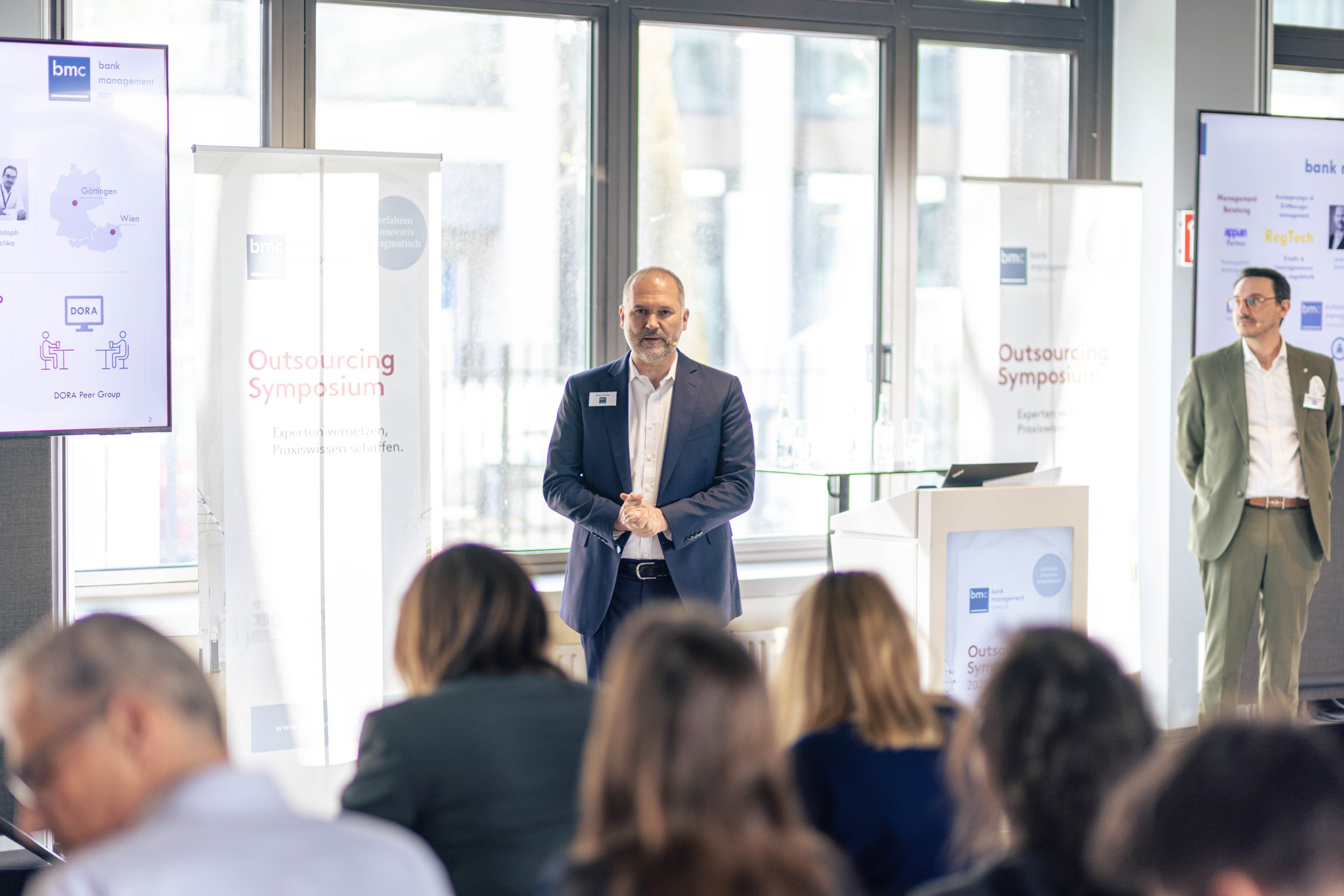 Ein Mann in Anzug steht auf einer Bühne bei einer Konferenz mit einem Mikrofon in der Hand, während ein anderer Mann im Hintergrund auf der Bühne steht. Es gibt eine Bildschirmwand mit Präsentationsfolien und Banner mit der Aufschrift "Outsourcing Sy