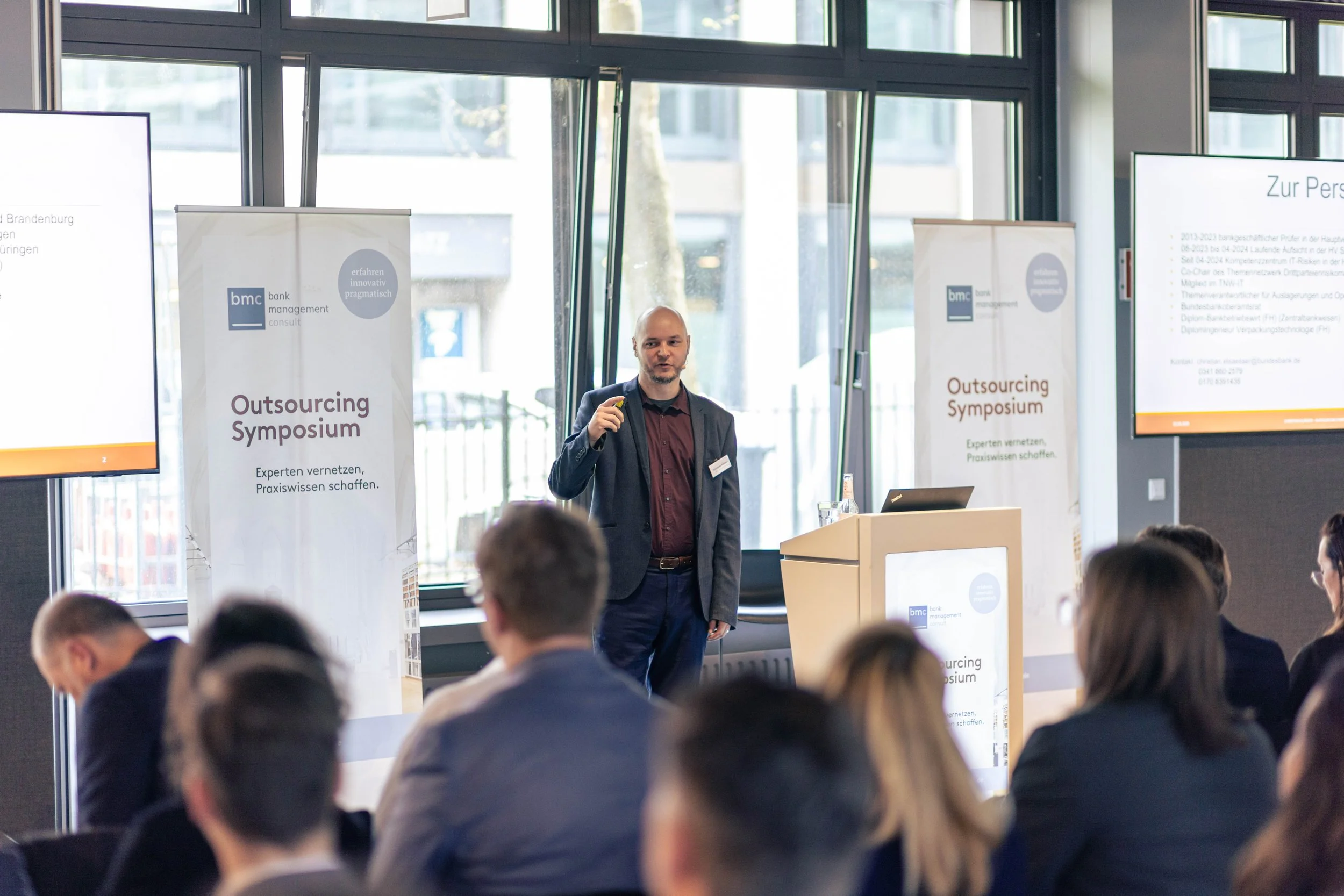 Ein Mann hält eine Präsentation beim Outsourcing-Symposium vor einem Publikum, mit zwei großen Monitoren und Bannern im Hintergrund.