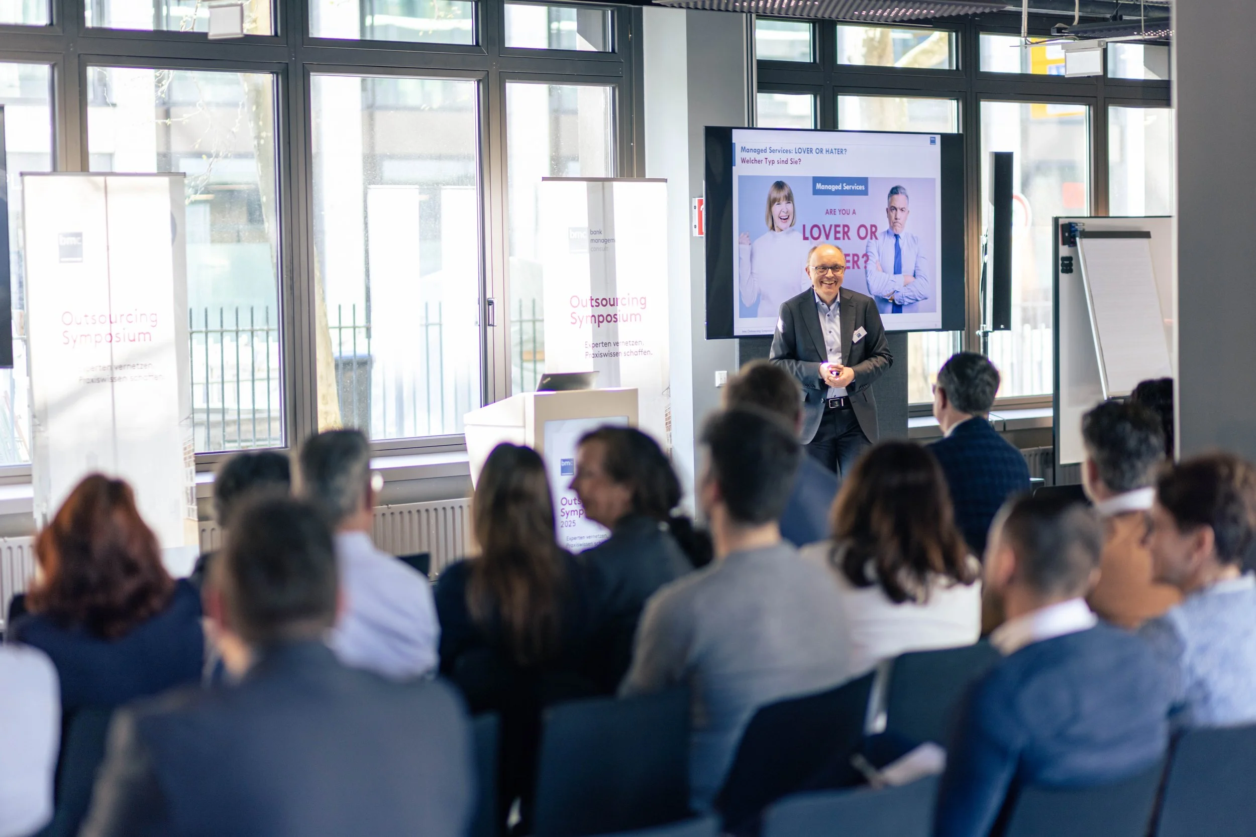 Ein Mann hält einen Vortrag vor einem Publikum während eines Outsourcing-Symposiums. Im Hintergrund ein Bildschirm mit einer Präsentation, außerdem stehen Banner und ein Whiteboard.
