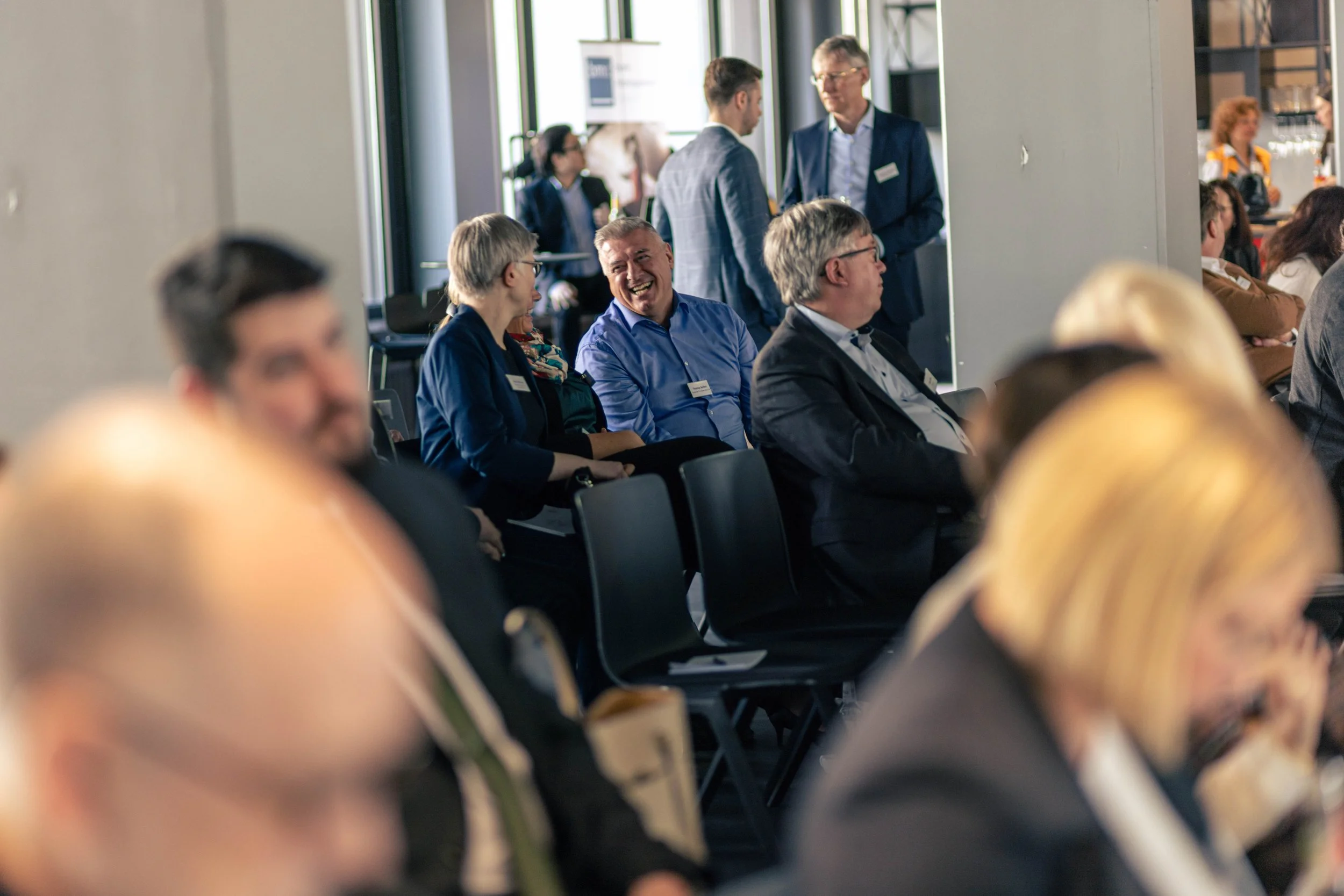 Menschen bei einer Konferenz oder einem Treffen, einige sitzen und lachen, andere stehen im Hintergrund auf einem modernen Veranstaltungsraum.