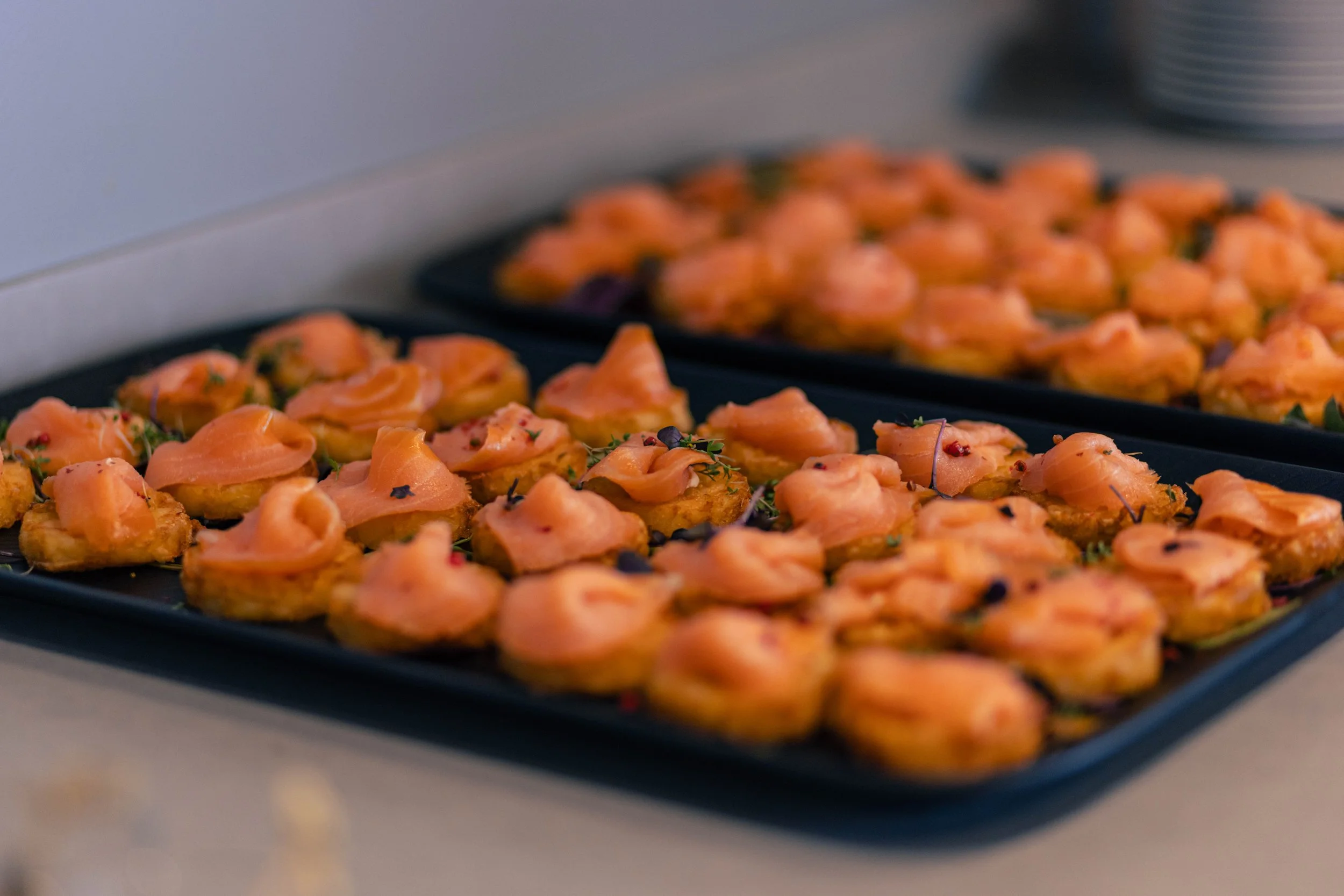 Zwei schwarze Platten mit kleinen, belegten Broten, Lachs und Kräuter, auf einem weißen Tisch, in der Nähe eines Kühlgeräts.