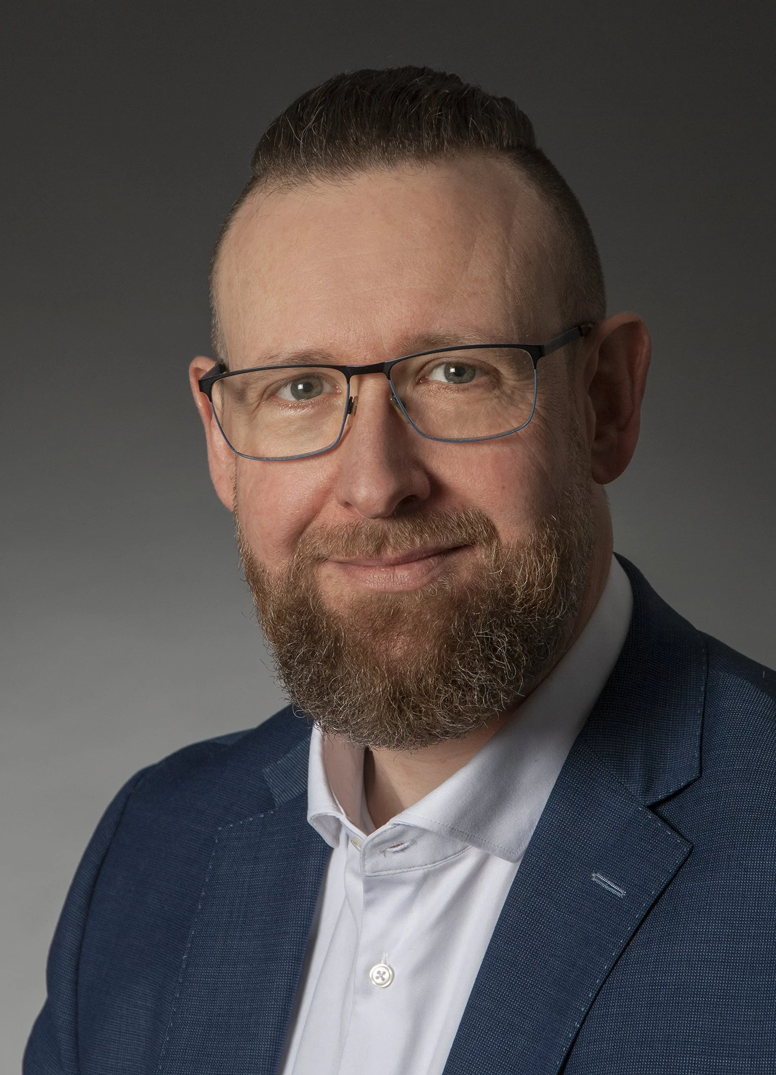Porträt eines Mannes mit Brille, braunem Bart und kurzem Haar, trägt einen dunkelblauen Anzug und ein weißes Hemd, vor grauem Hintergrund.