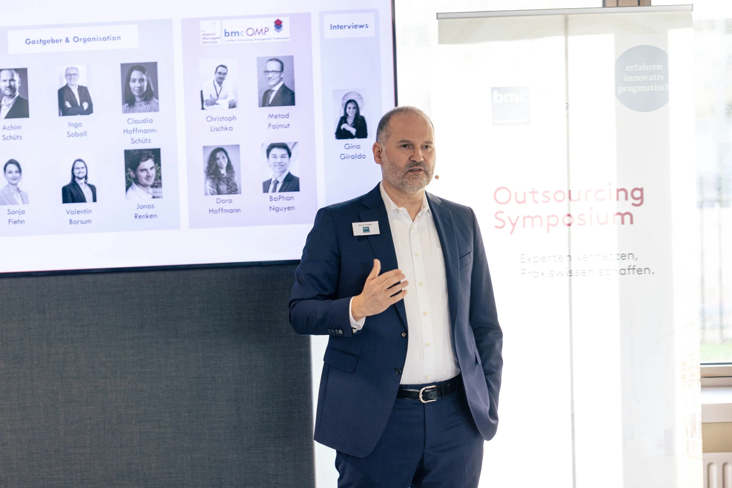 Mann in dunklem Anzug beim Outsourcing-Symposium spricht vor großem Bildschirm mit Fotos und Namen von Personen sowie Texten.